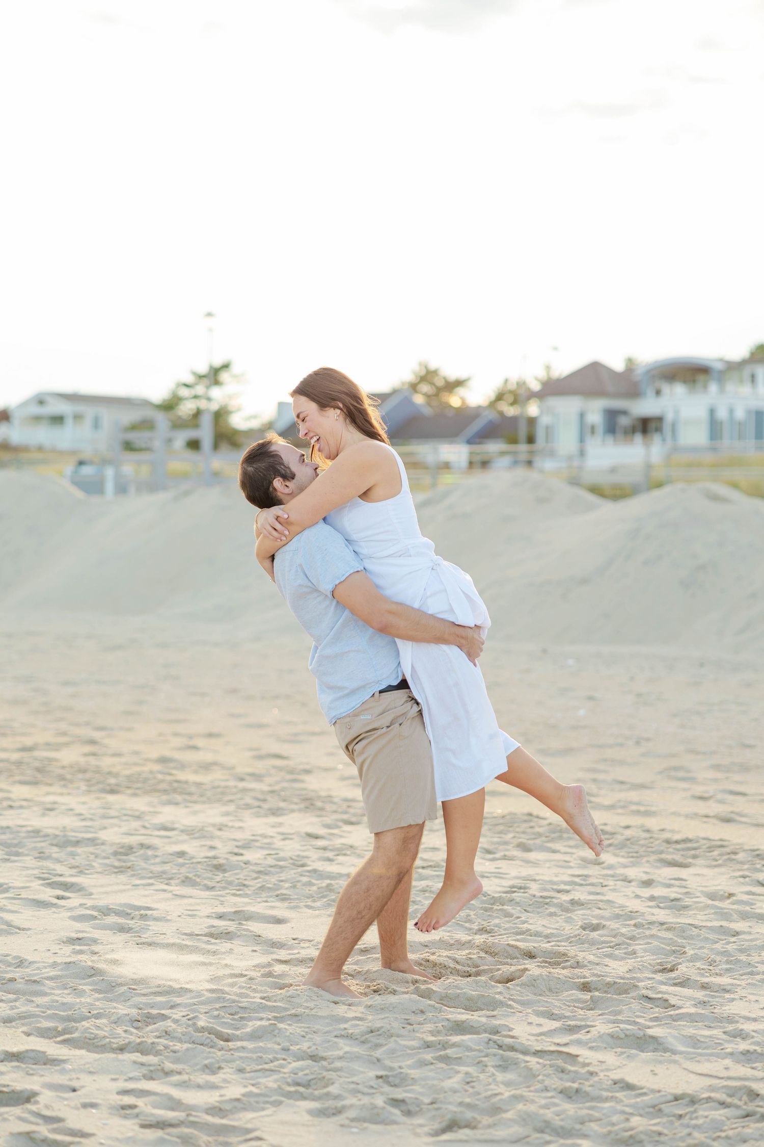 A couple embraces playfully on a sandy beach at sunset with beach houses visible in the background.