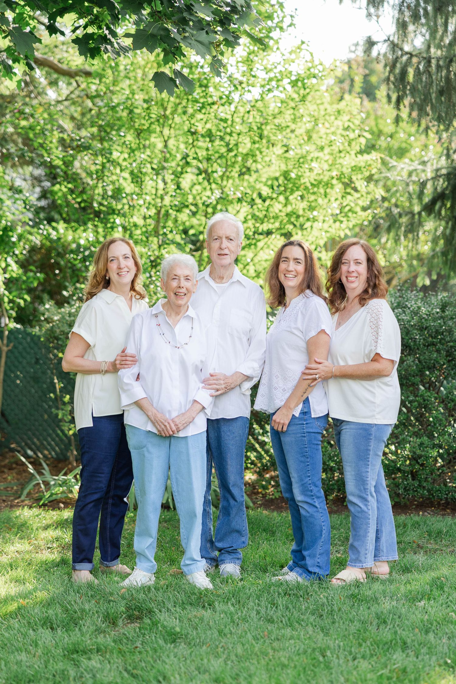 Family members wearing matching white shirts and blue jeans pose together in a sunny garden setting.