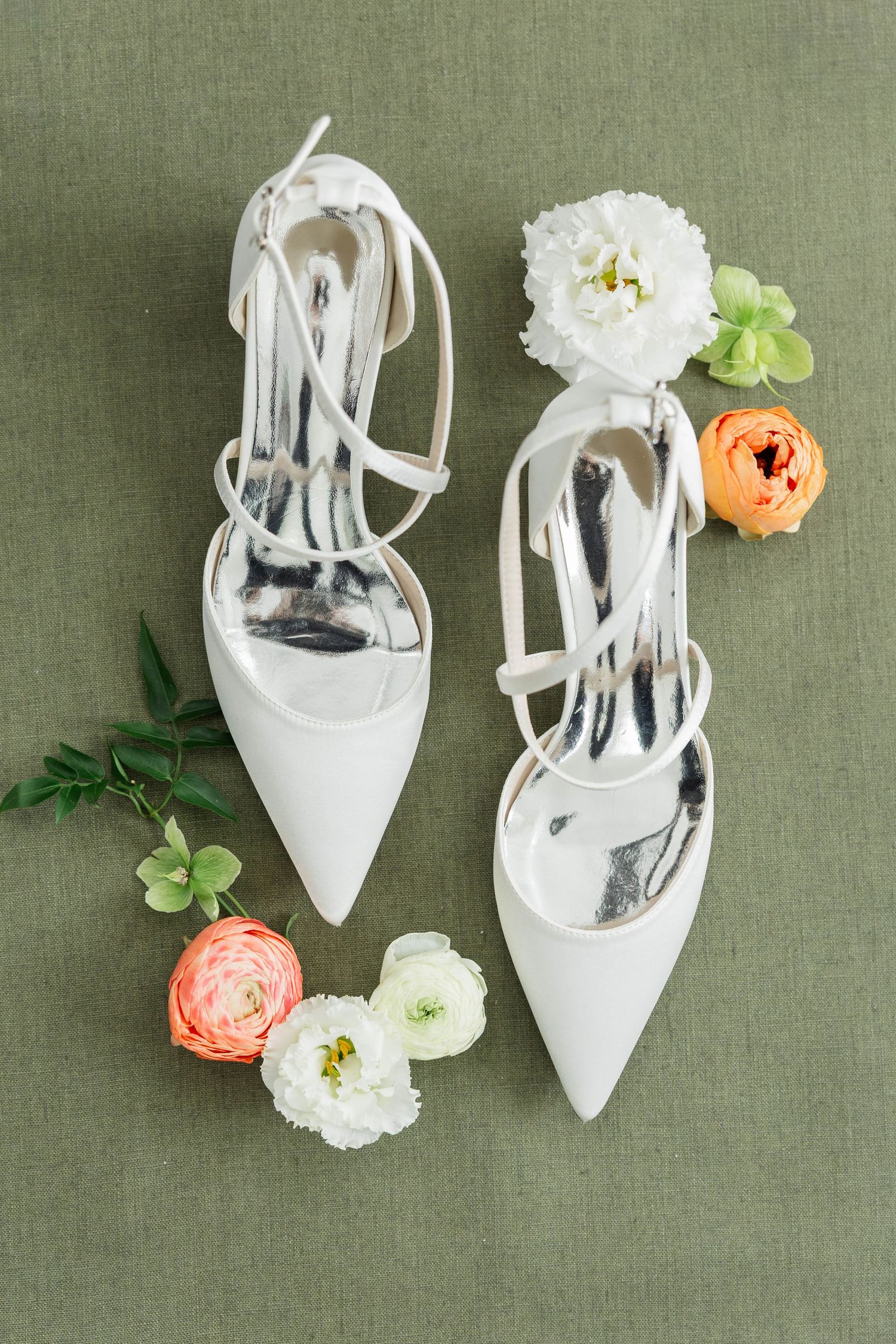 White bridal heels with ankle straps surrounded by peach and white ranunculus flowers on sage green linen.