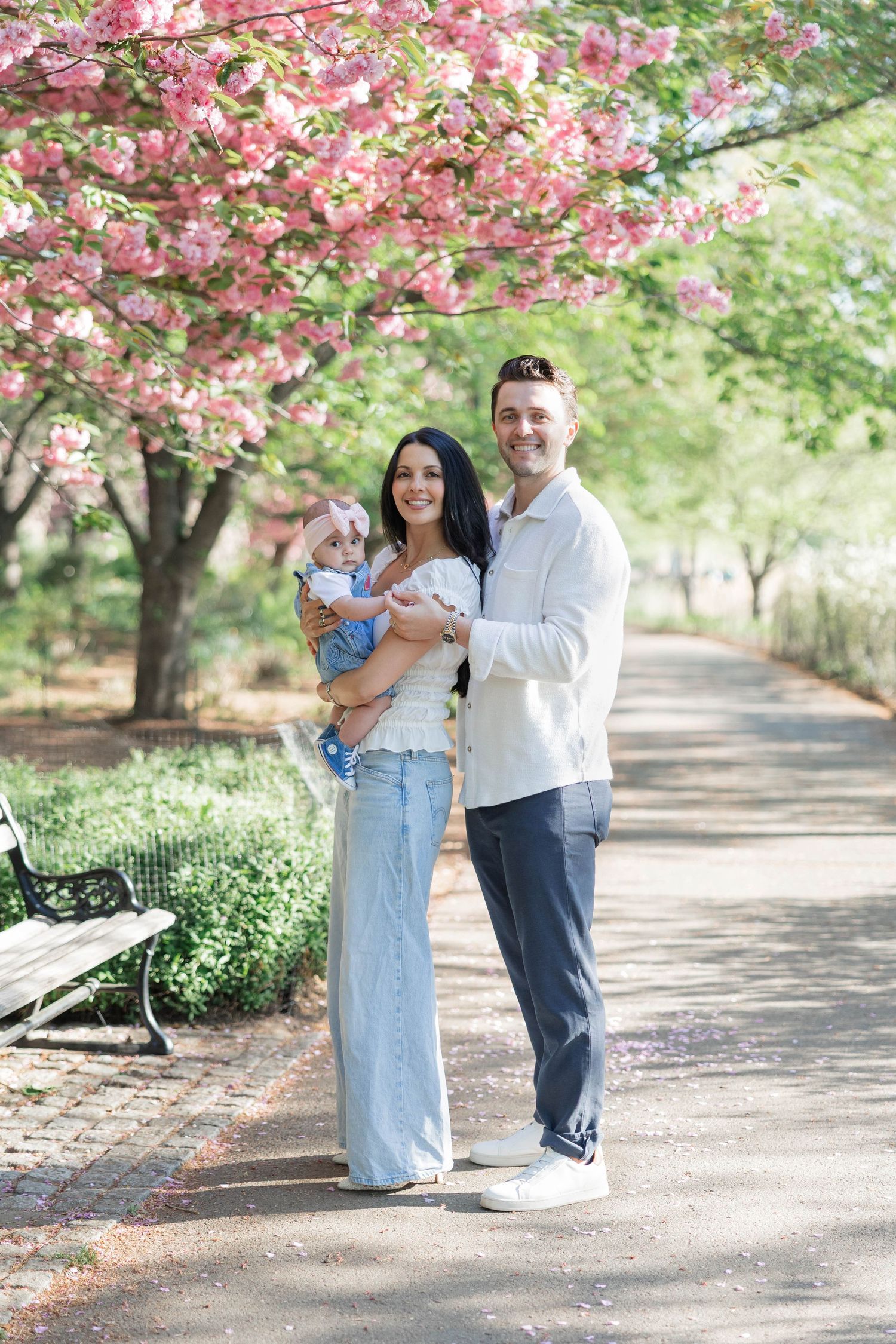 A happy family poses under pink cherry blossom trees on a park path, parents holding their baby girl in spring.