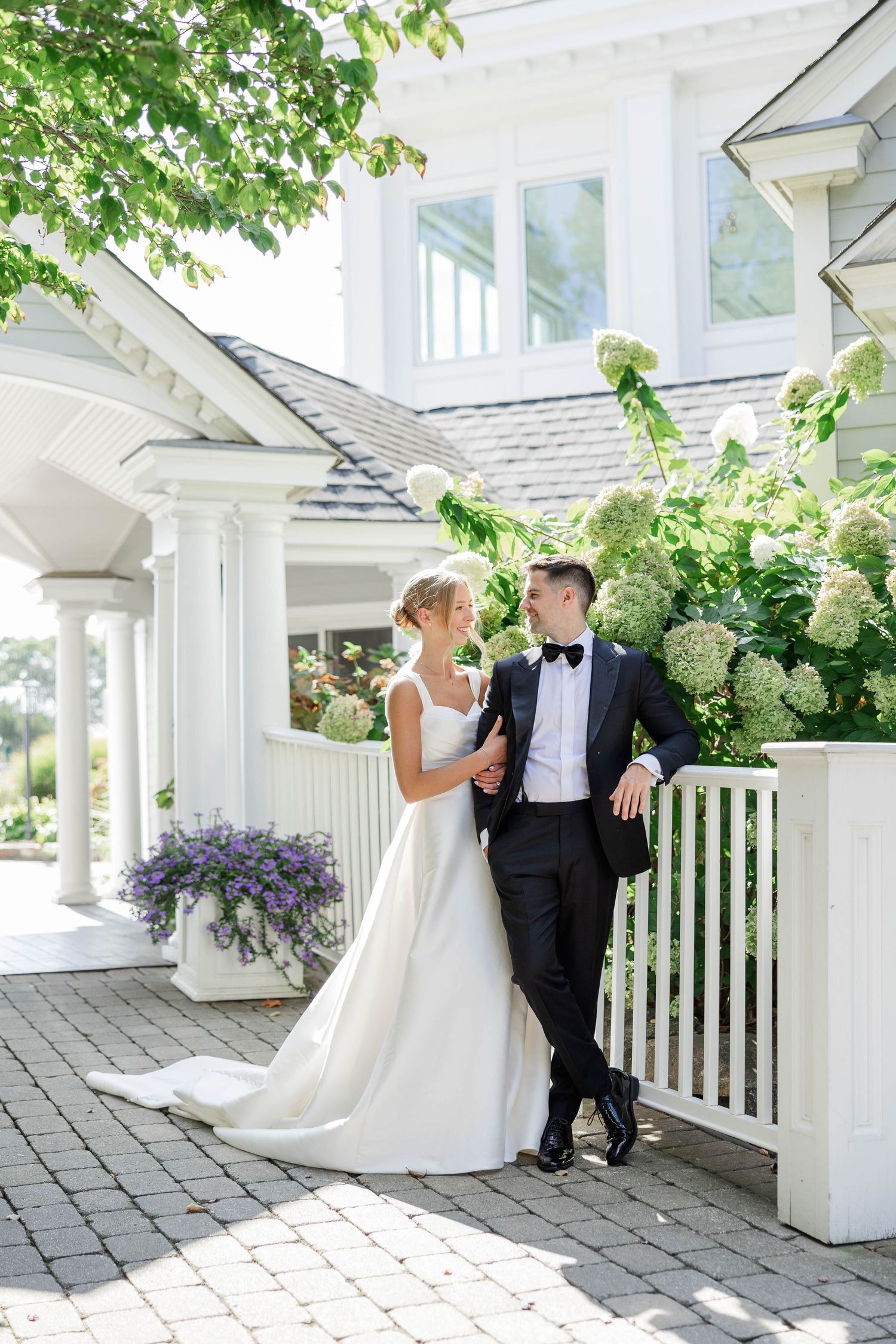 A romantic wedding couple shares intimate moments under a white pergola surrounded by lush greenery and hydrangeas.