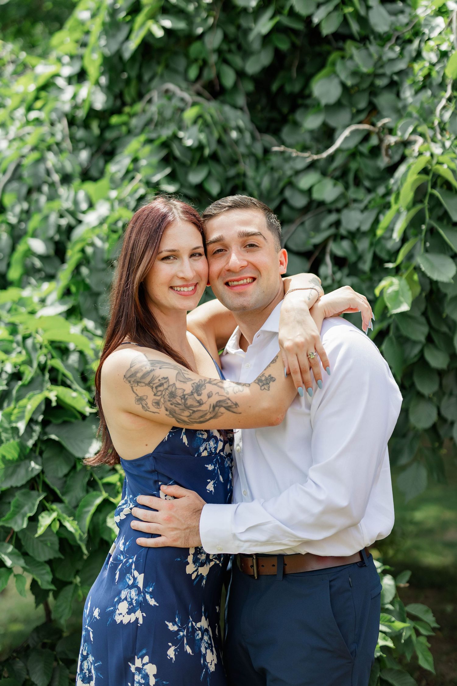 A smiling couple embraces outdoors, woman in floral blue dress with tattoos hugs man in white shirt amid lush green leaves.
