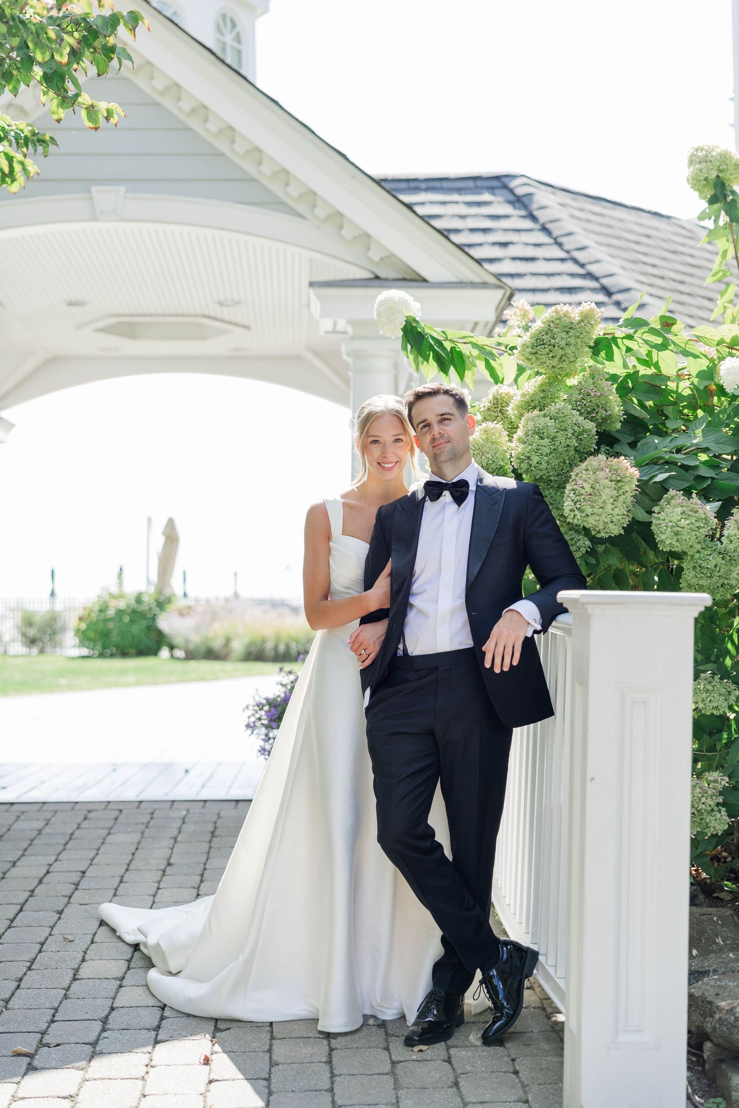 A couple in wedding attire sits together on white porch steps during an outdoor photoshoot.
