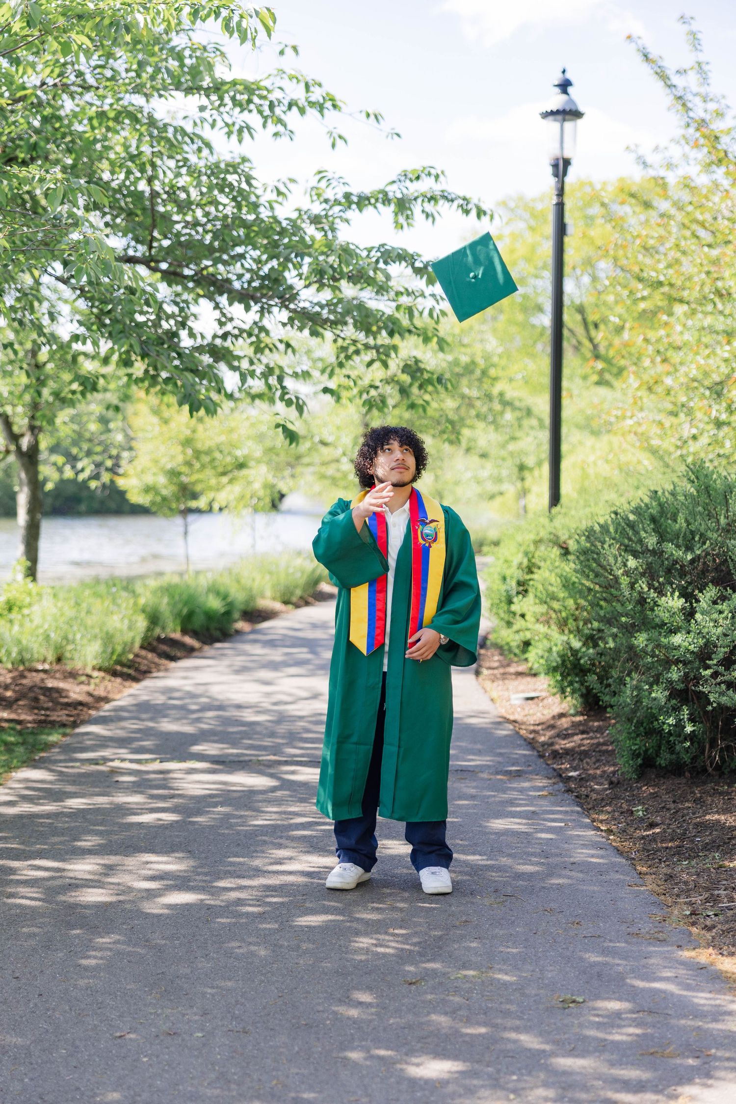 Graduate in green cap and gown tosses mortarboard into the air on a tree-lined park pathway on graduation day.