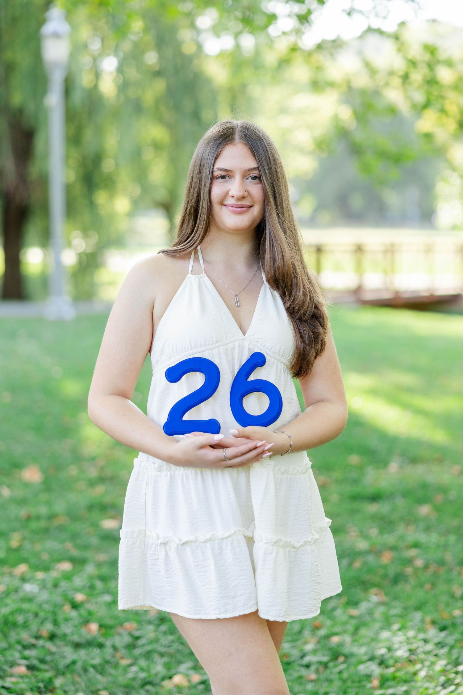 Young woman in white dress holding number 26 sign, smiling outdoors in green park setting.