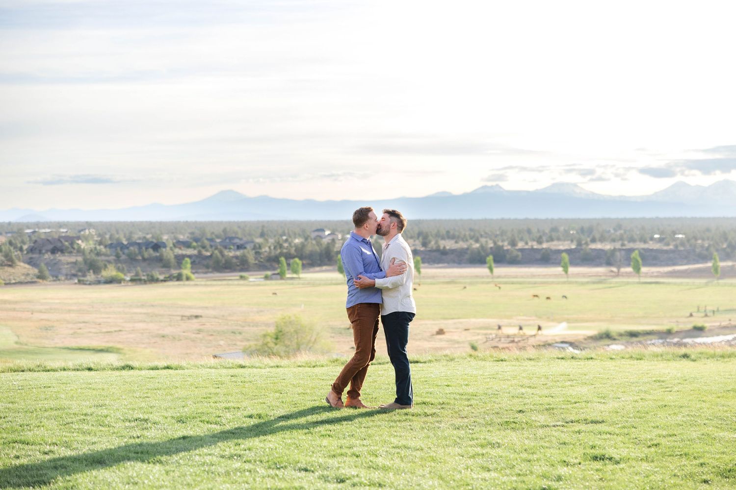 Two men share a romantic kiss on a lush green lawn with mountain views and open landscape in the background.