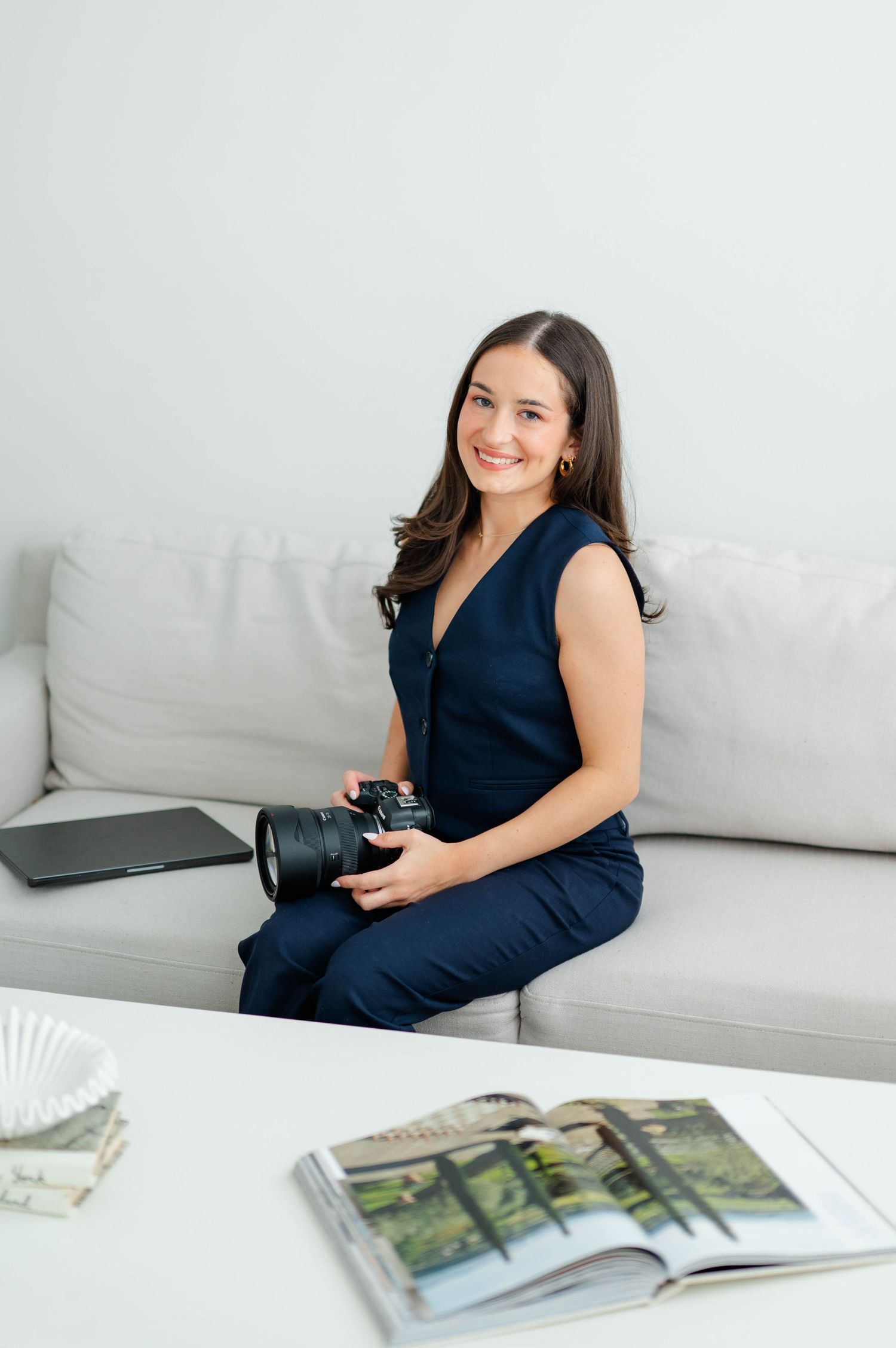 A person in a navy sleeveless top relaxes on a white couch while looking through a magazine.
