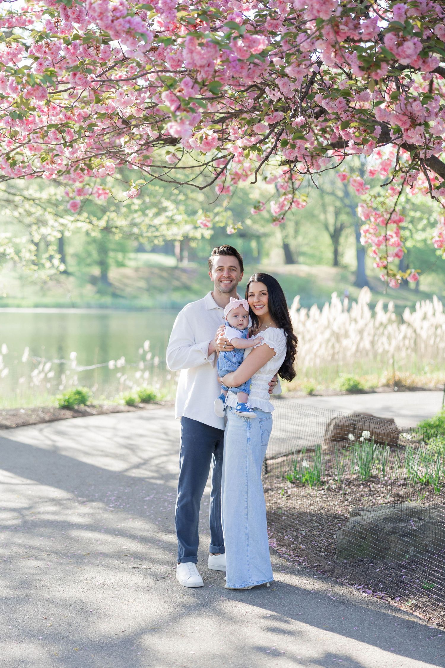 A couple holds their baby under pink cherry blossoms by a lake in spring.