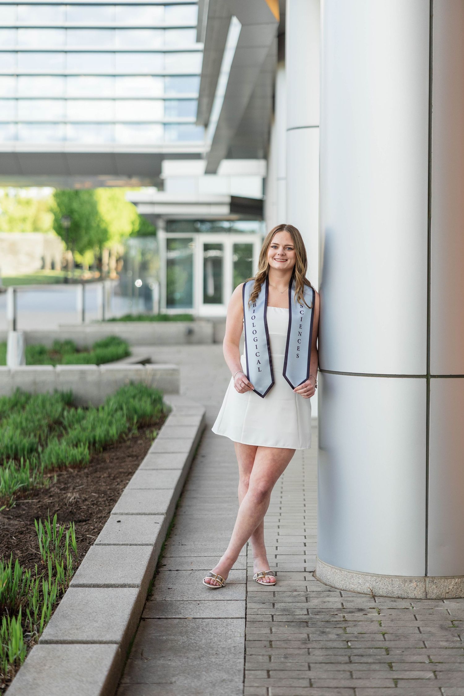Young woman in white dress and graduation stole leaning against a column outdoors at her graduation celebration.
