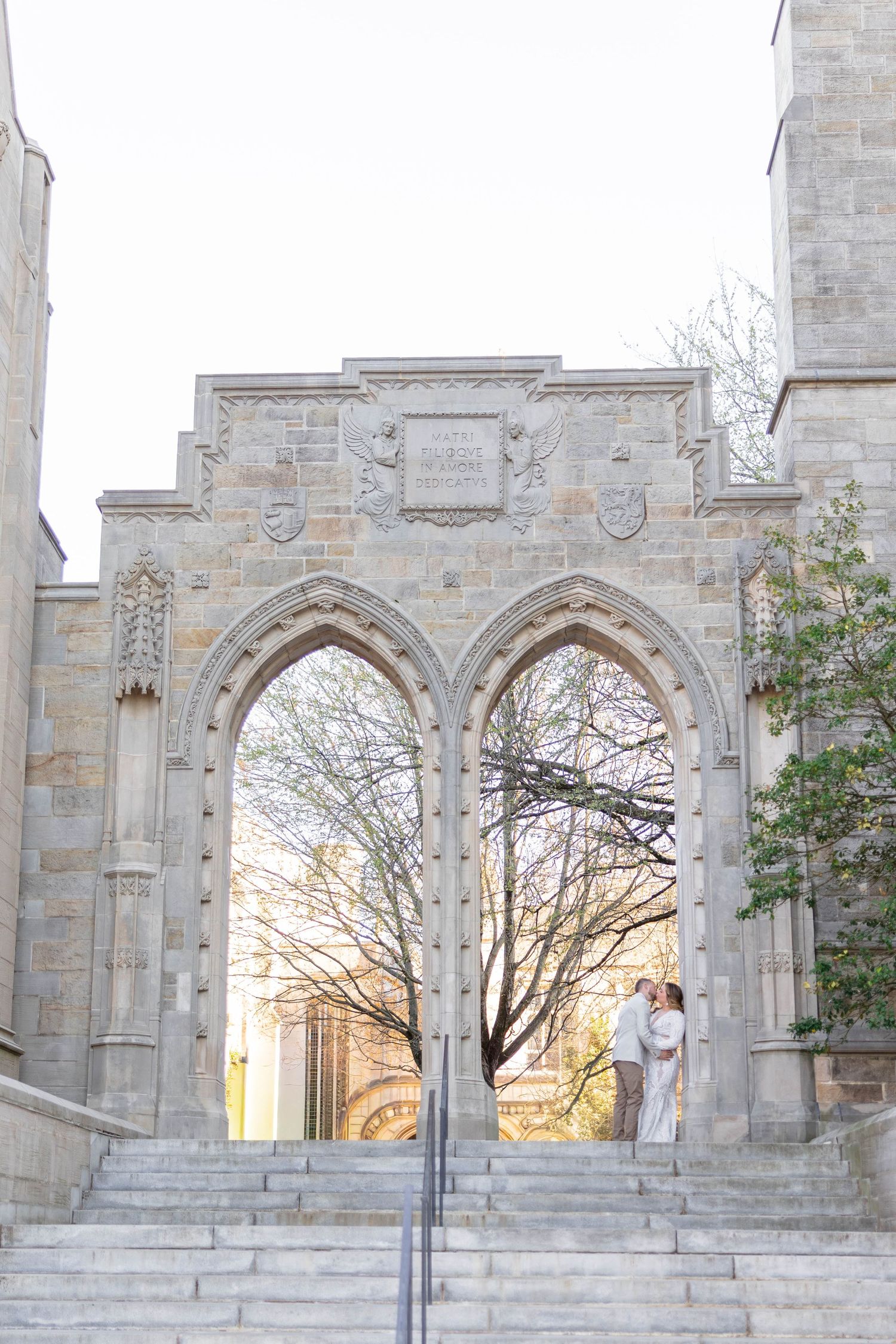 Couple embracing under Gothic stone arches on university campus steps, framed by ornate carved stonework and bare trees.