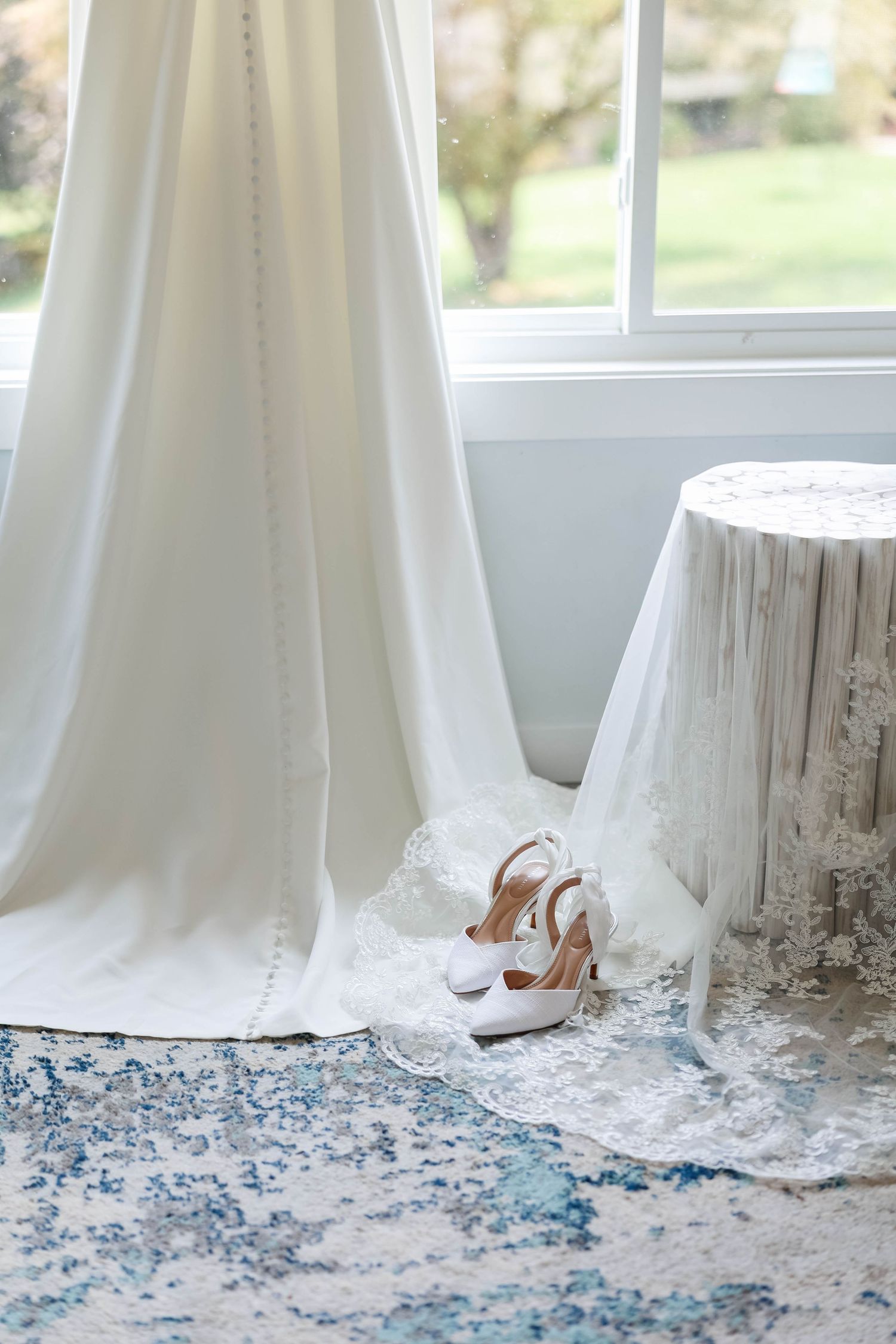 White wedding dress with lace hem displayed near window with bridal heels on blue patterned rug.