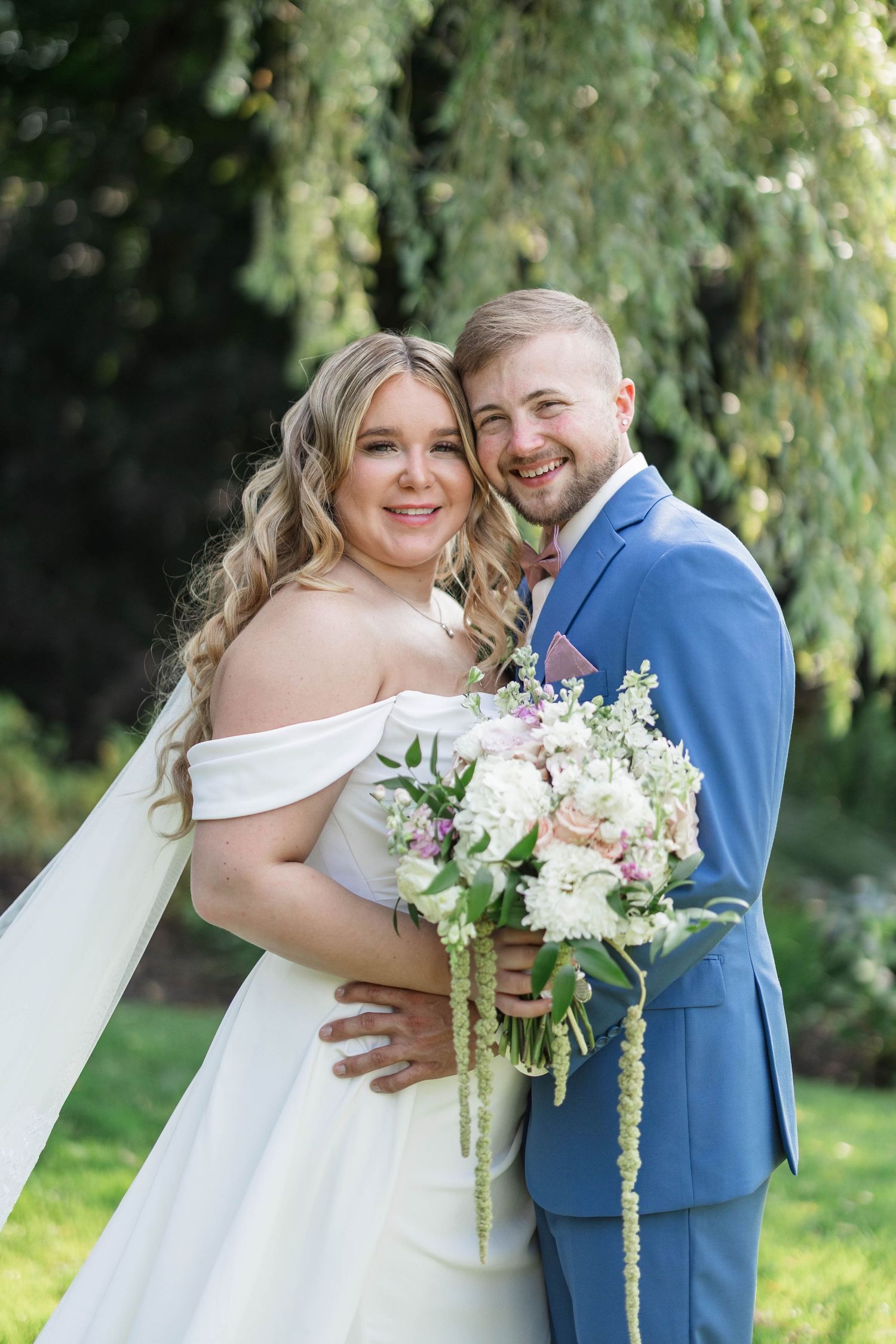 Smiling bride in off-shoulder gown holding bouquet with groom in blue suit, posing outdoors under a weeping willow tree.