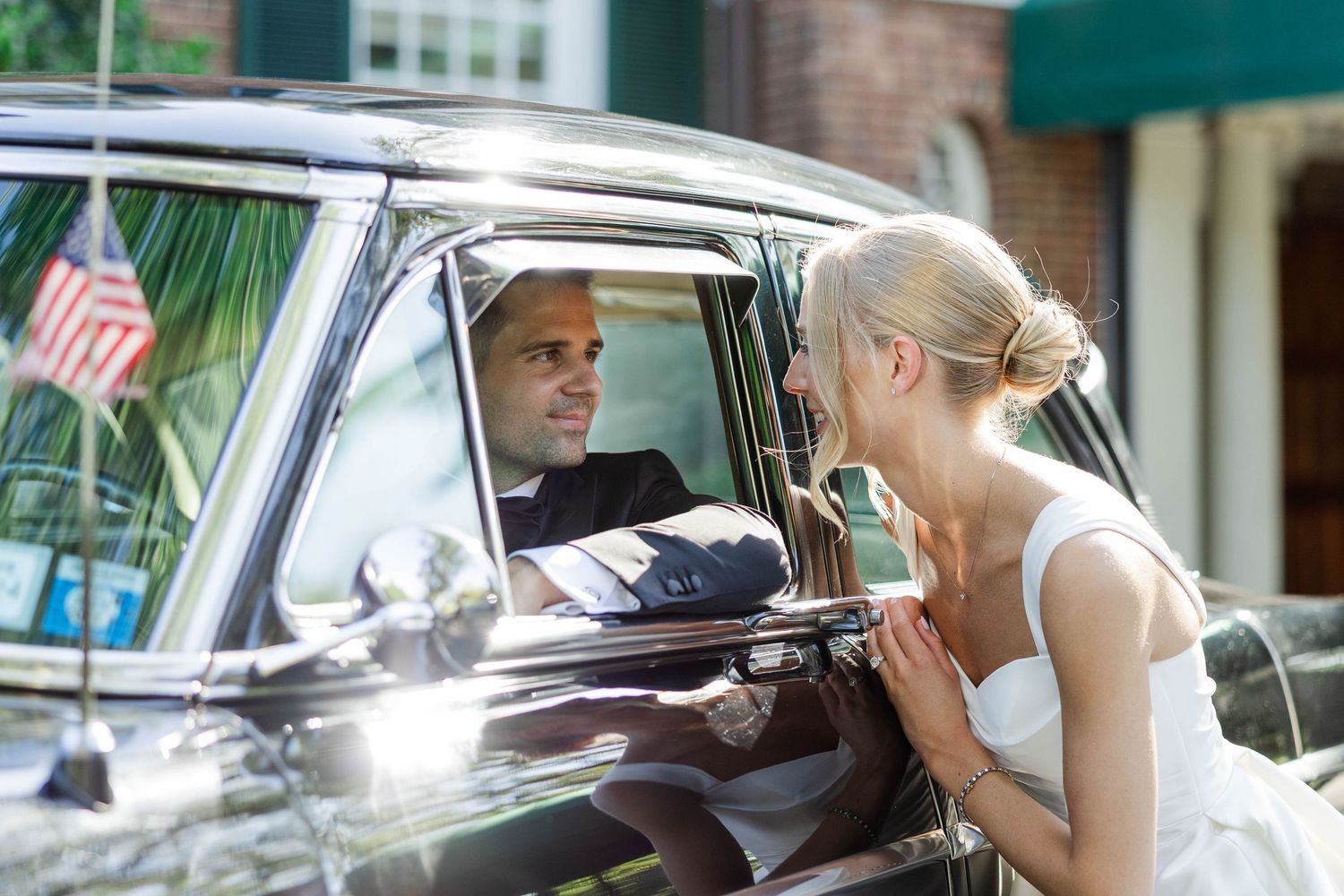 A couple shares a romantic moment through the window of a classic black car with an American flag visible in the background.