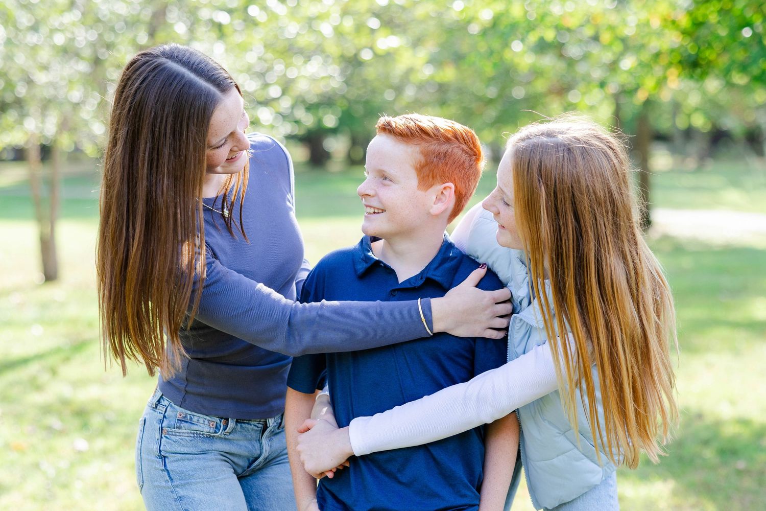 Three people laughing and hugging while enjoying a fun moment together outdoors in a park setting.