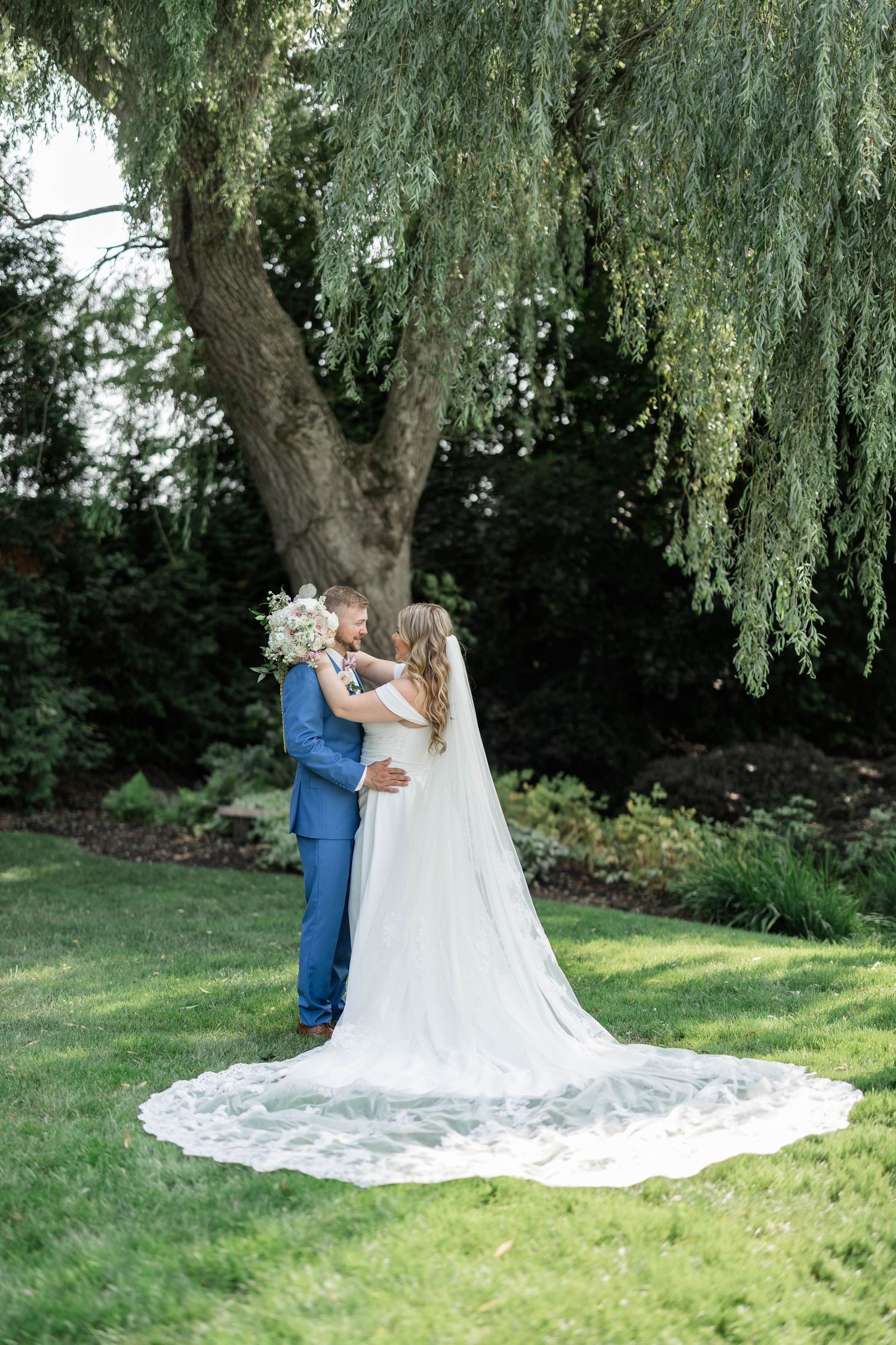 Bride and groom embrace under a weeping willow tree, bride wearing a long lace veil trailing on green grass.