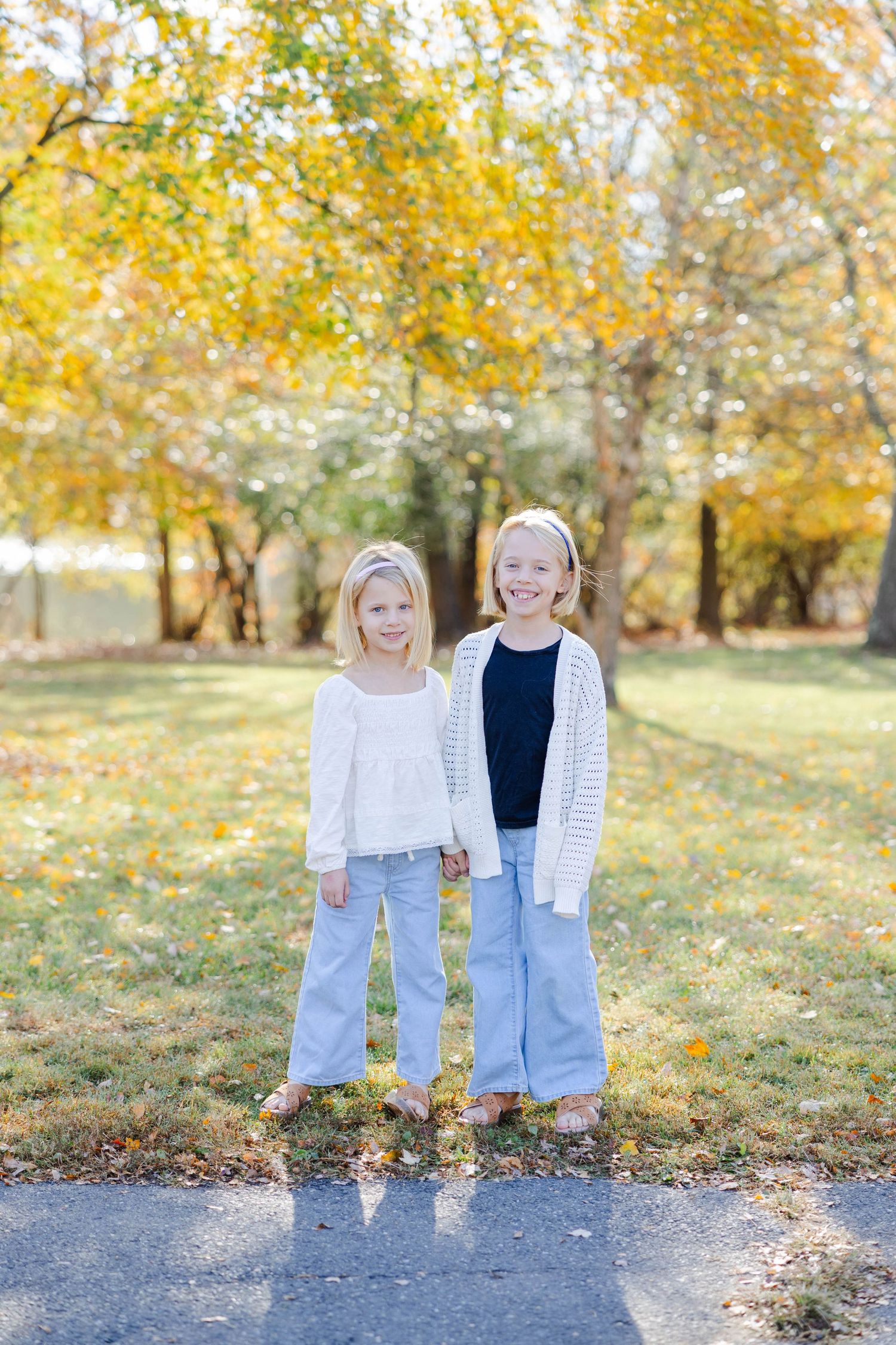 Two young children stand together in a park with golden autumn leaves in the background.