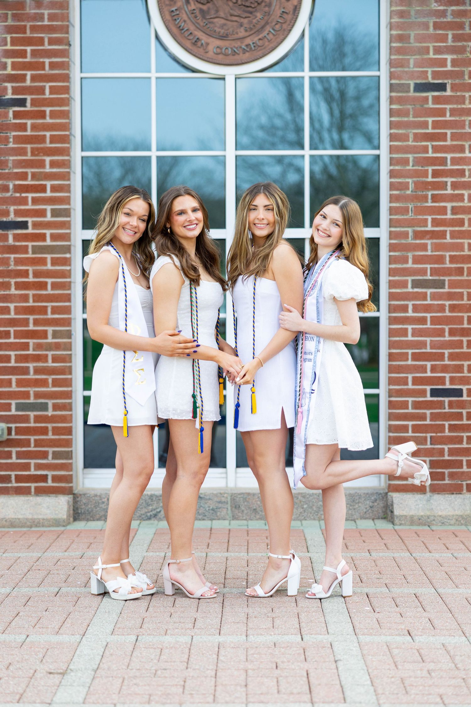 Four young women in white dresses and graduation stoles pose together in front of a brick building.