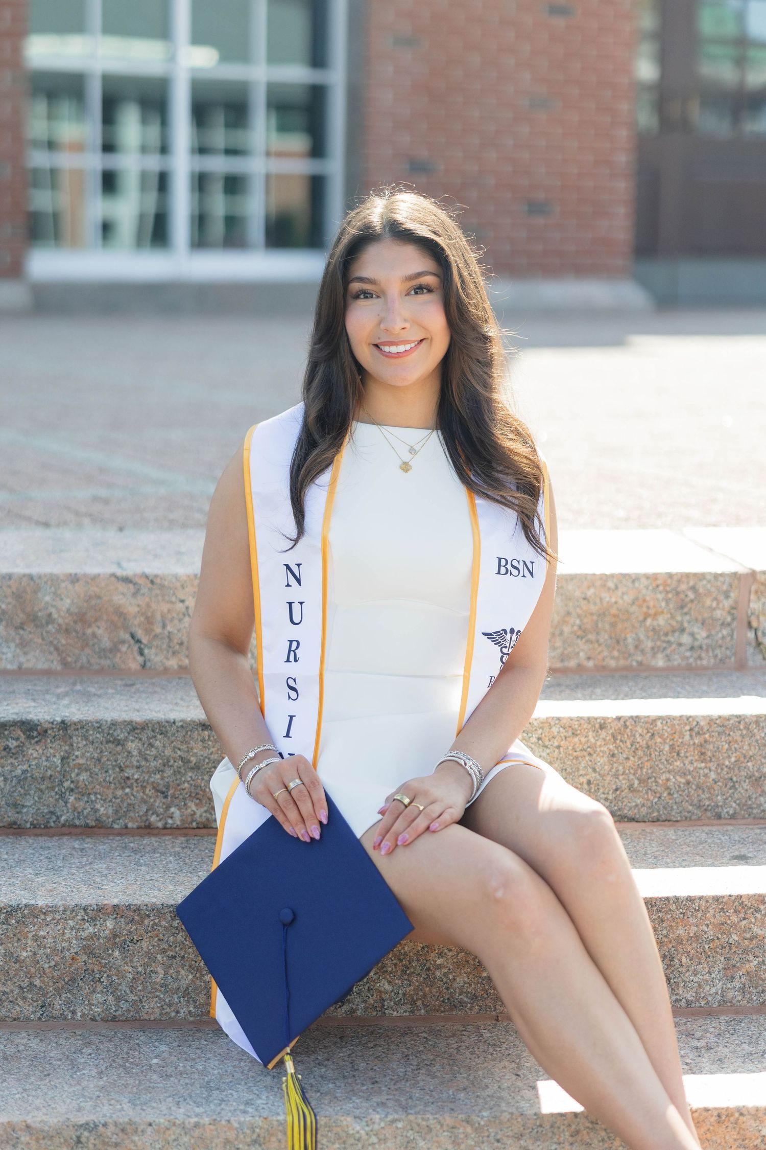 Smiling nursing graduate in white dress and BSN sash sits on steps holding navy graduation cap with gold tassel.