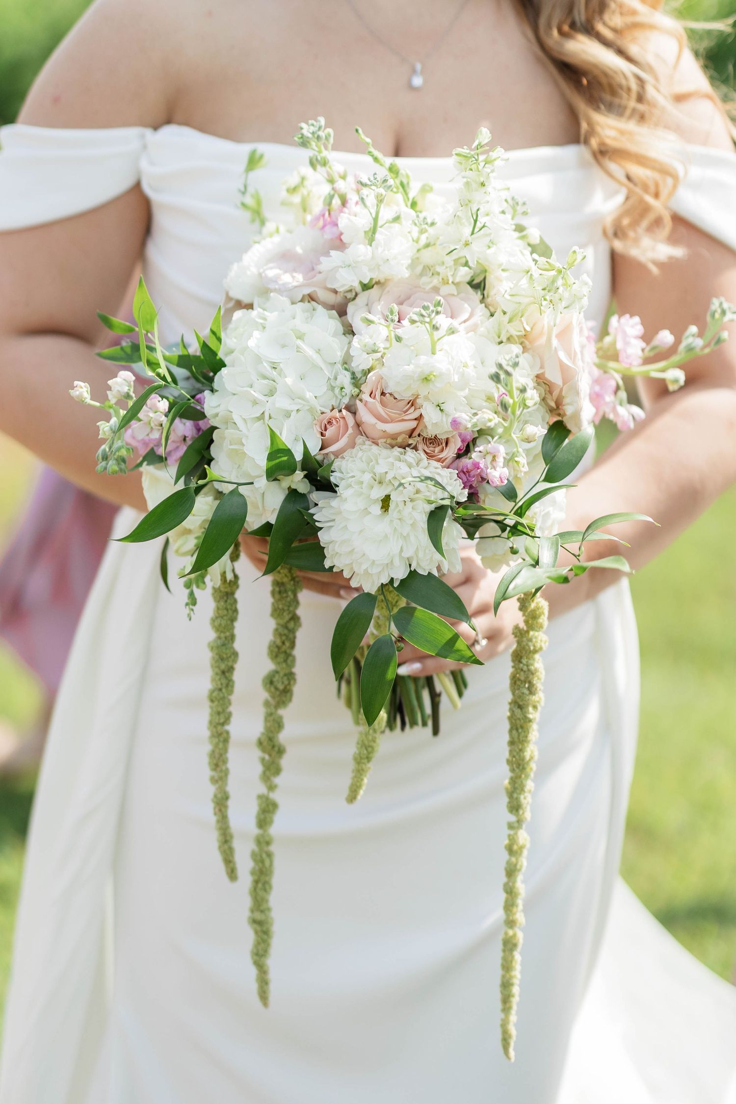 Bride holding a lush wedding bouquet with white, blush pink flowers and cascading green amaranthus in outdoor setting.