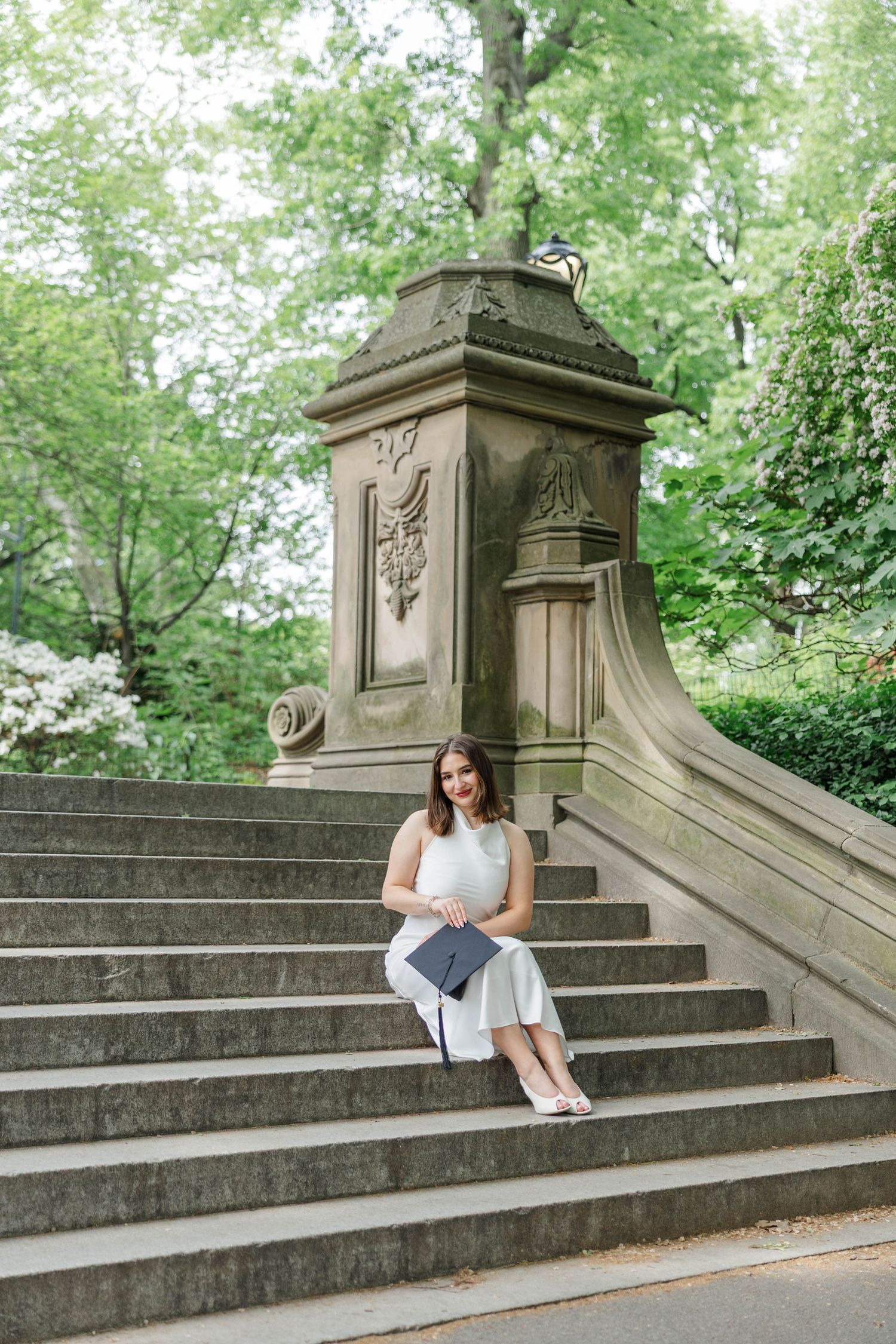 A woman in a white dress sits on stone steps holding a graduation cap, surrounded by lush green trees in a park.