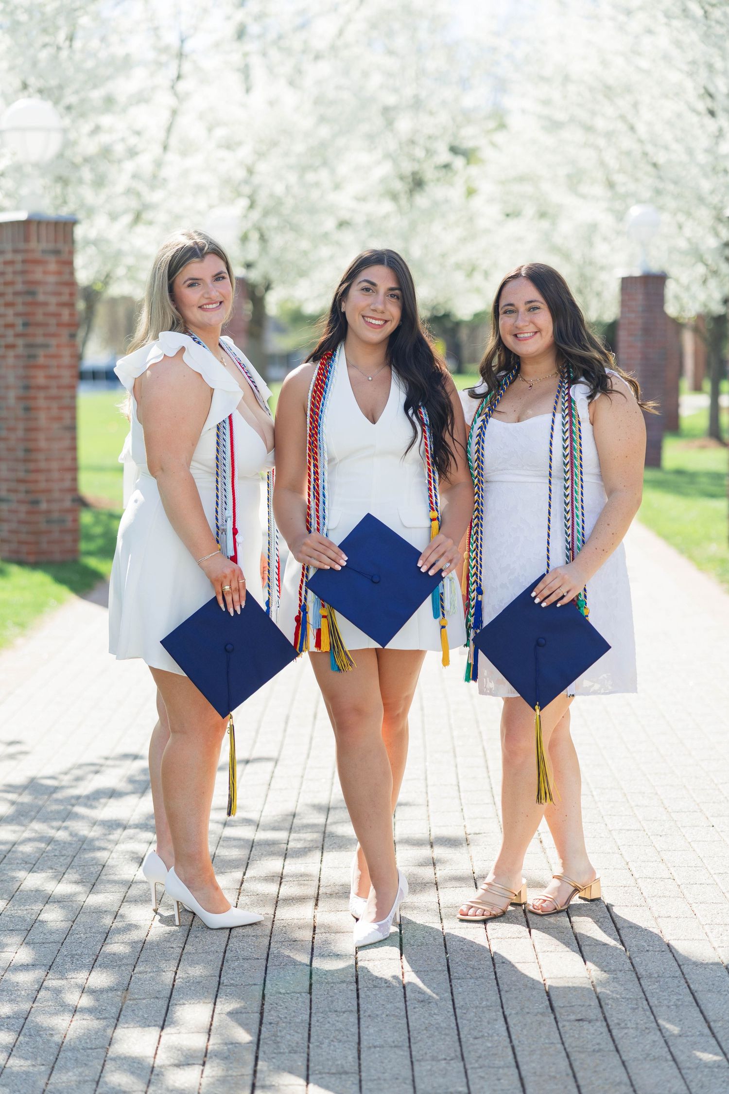 Three smiling women in white dresses hold blue graduation caps outdoors near blooming trees on a sunny day.