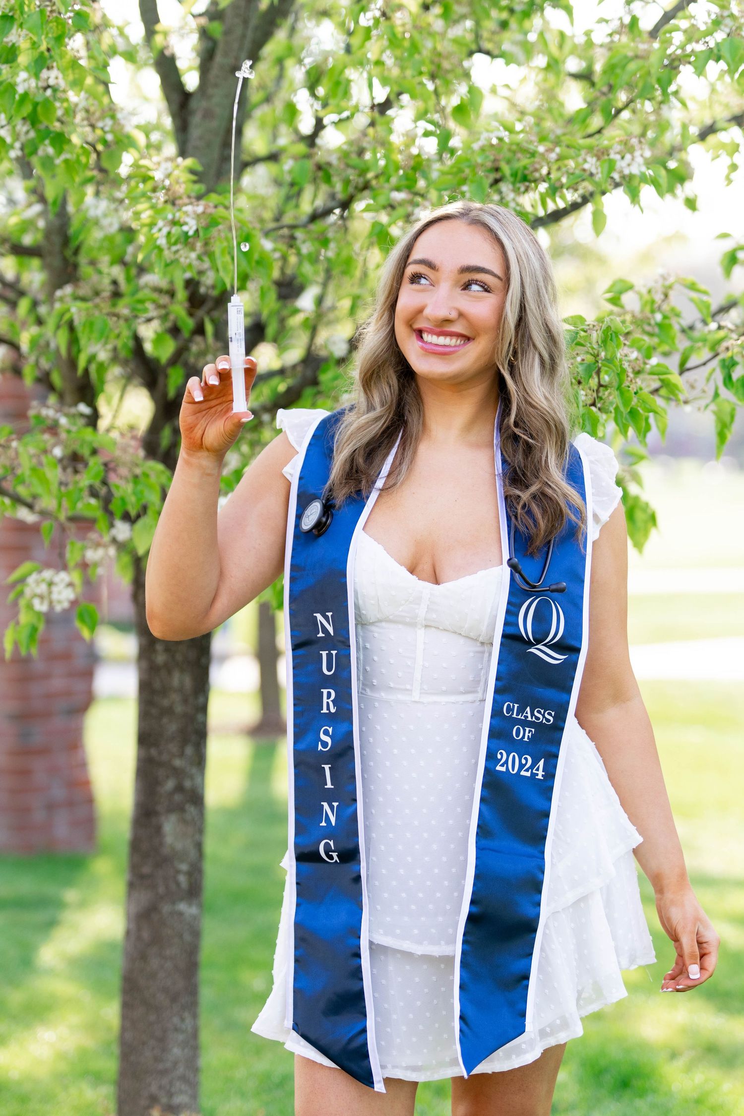 Nursing graduate in white dress and blue sash celebrating outdoors among trees.