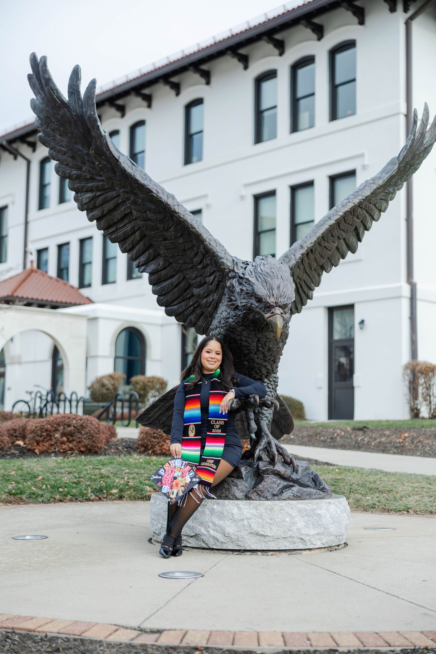 Graduate poses in colorful stole before large bronze eagle statue outside white university building.