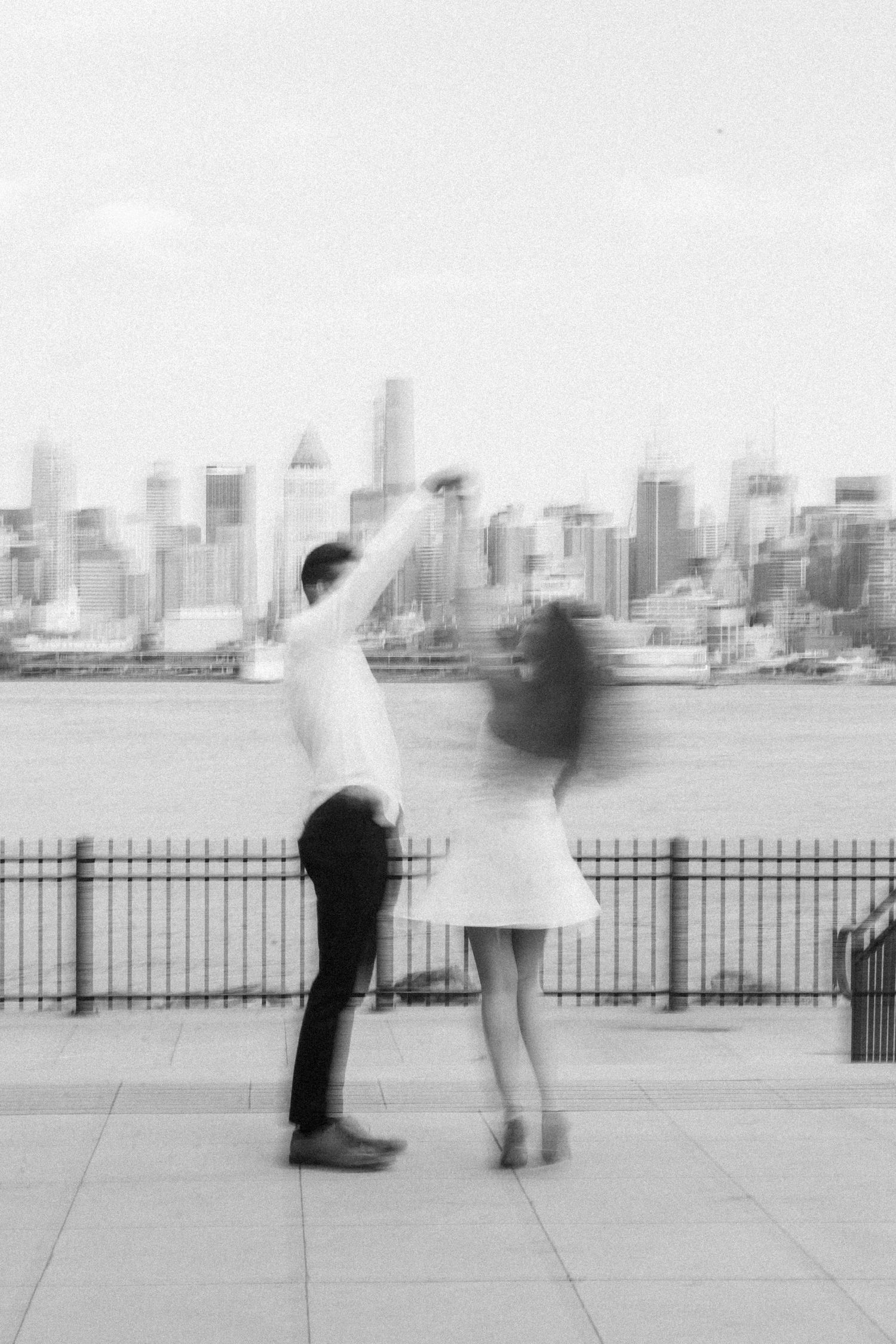 Blurred black and white motion portrait of people standing near a waterfront fence with a cityscape in the background.