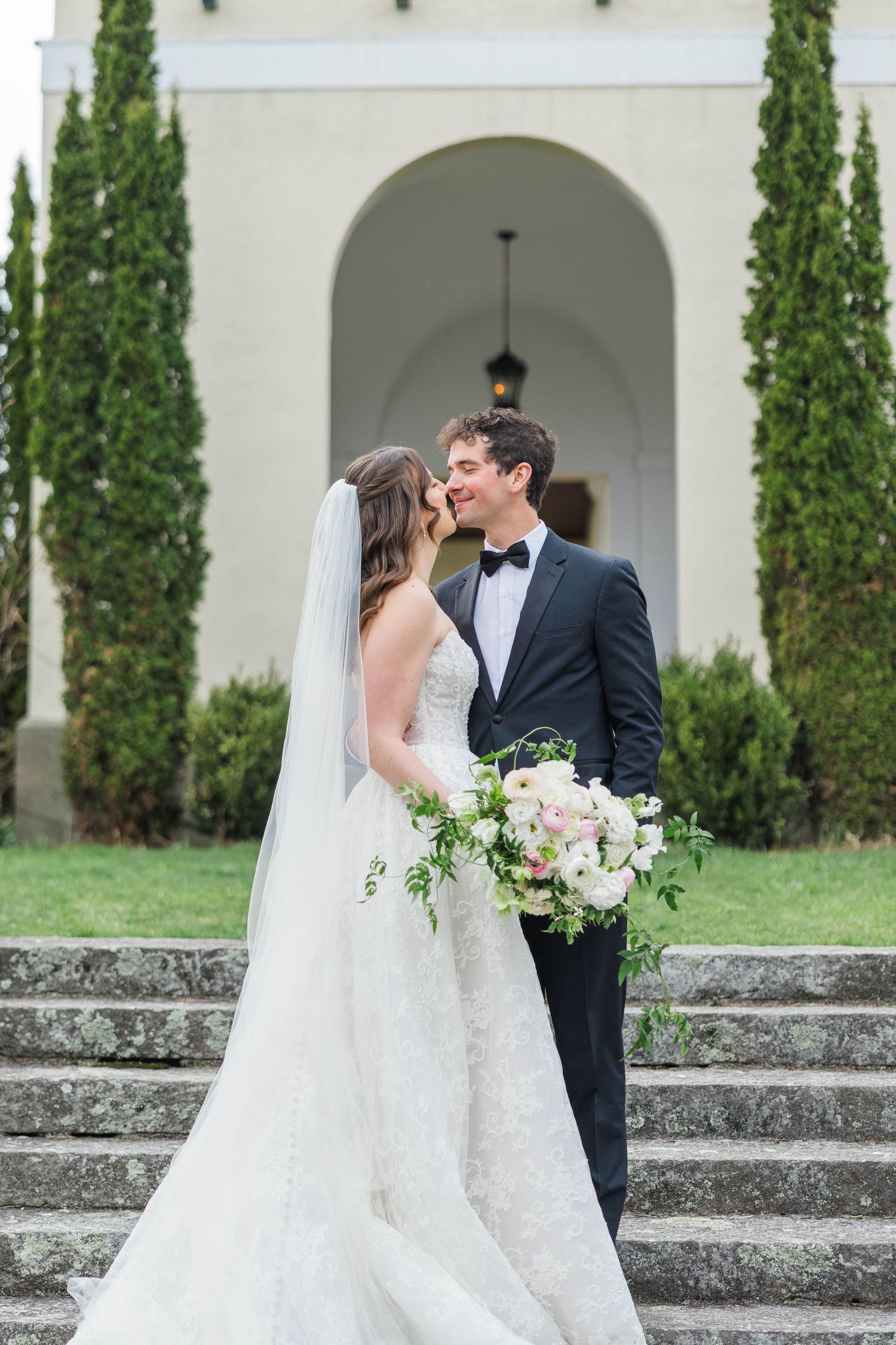 Bride and groom share a kiss on stone steps outside an elegant venue with arched doorway and tall cypress trees.