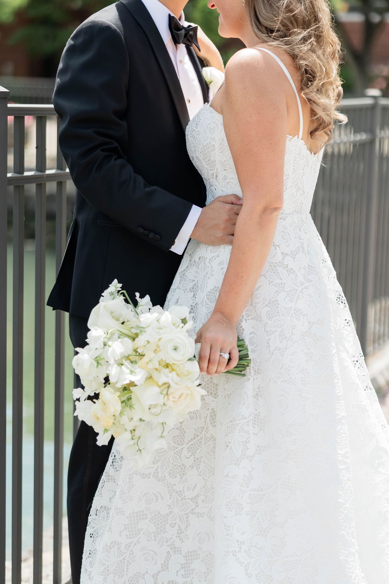 Bride in lace wedding gown holding white floral bouquet stands beside groom in black tuxedo outdoors.
