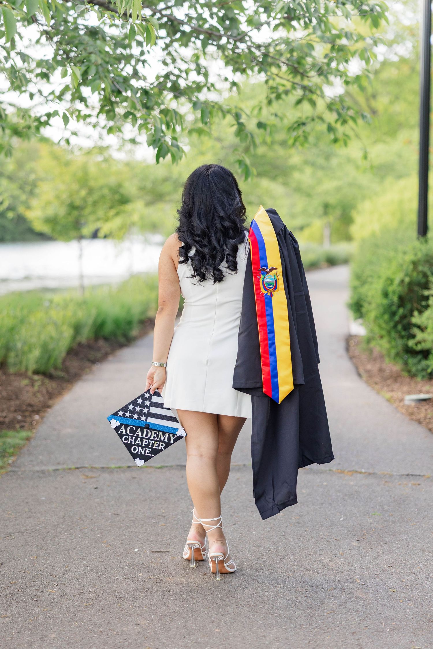 Graduate in white dress walks path holding cap and gown with colorful sash, celebrating academic achievement outdoors.