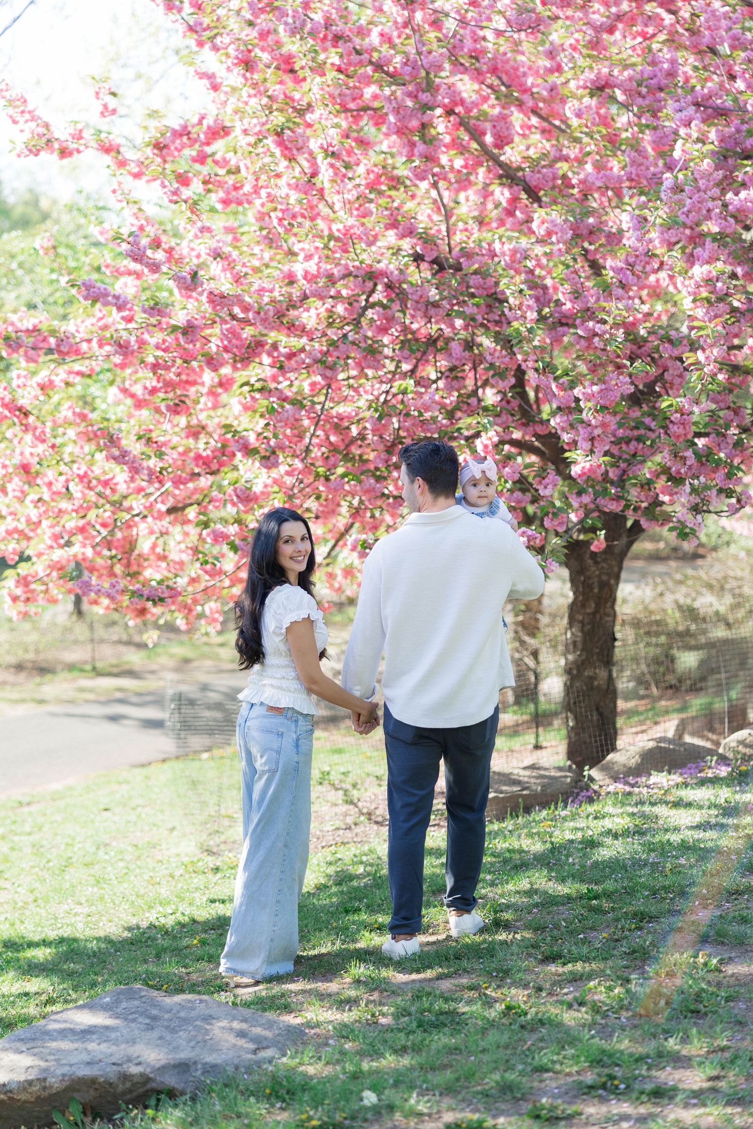A couple holding hands with their baby under blooming pink cherry blossom trees in a sunny park.