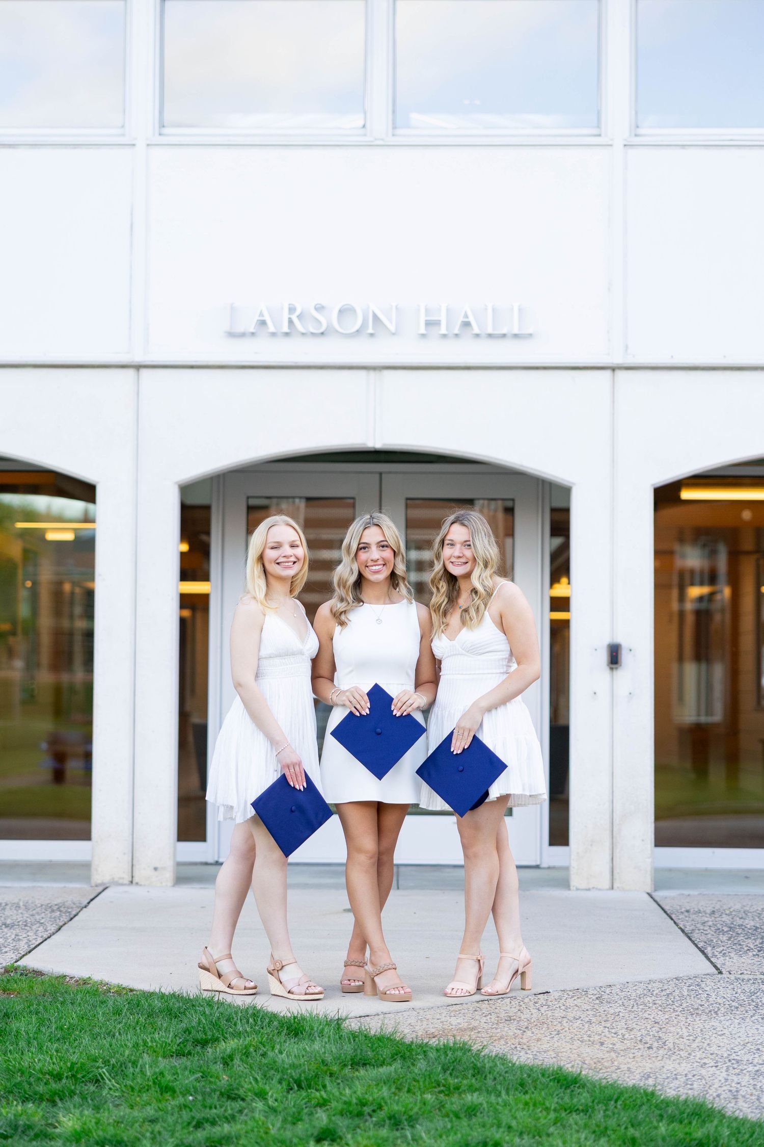 Three graduates hold blue caps while standing in front of Larson Hall entrance.