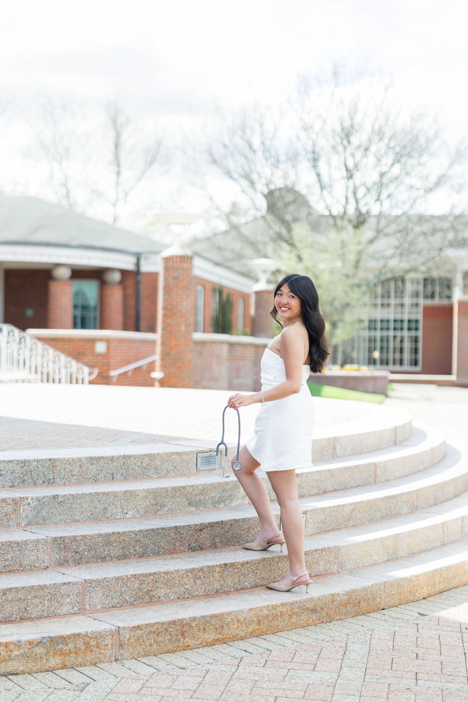Young woman in white dress holding purse, smiling on granite steps outside brick building.
