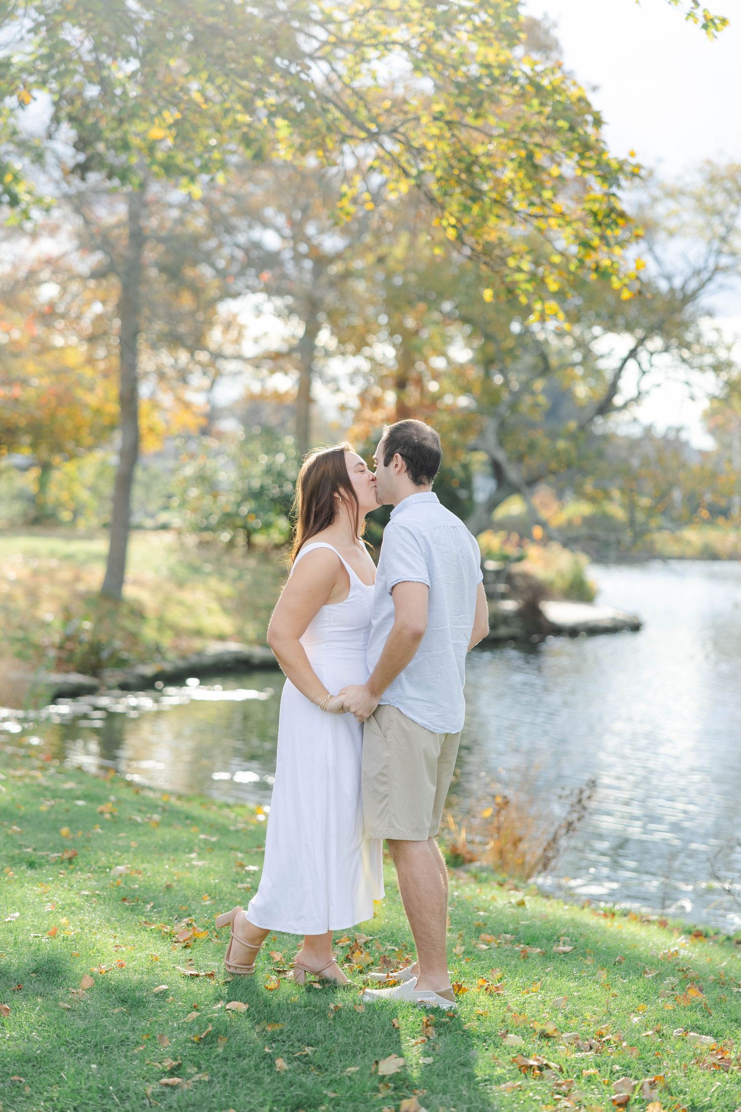 A romantic couple embraces by a tranquil lake on an autumn day with golden leaves and sunlight filtering through trees.