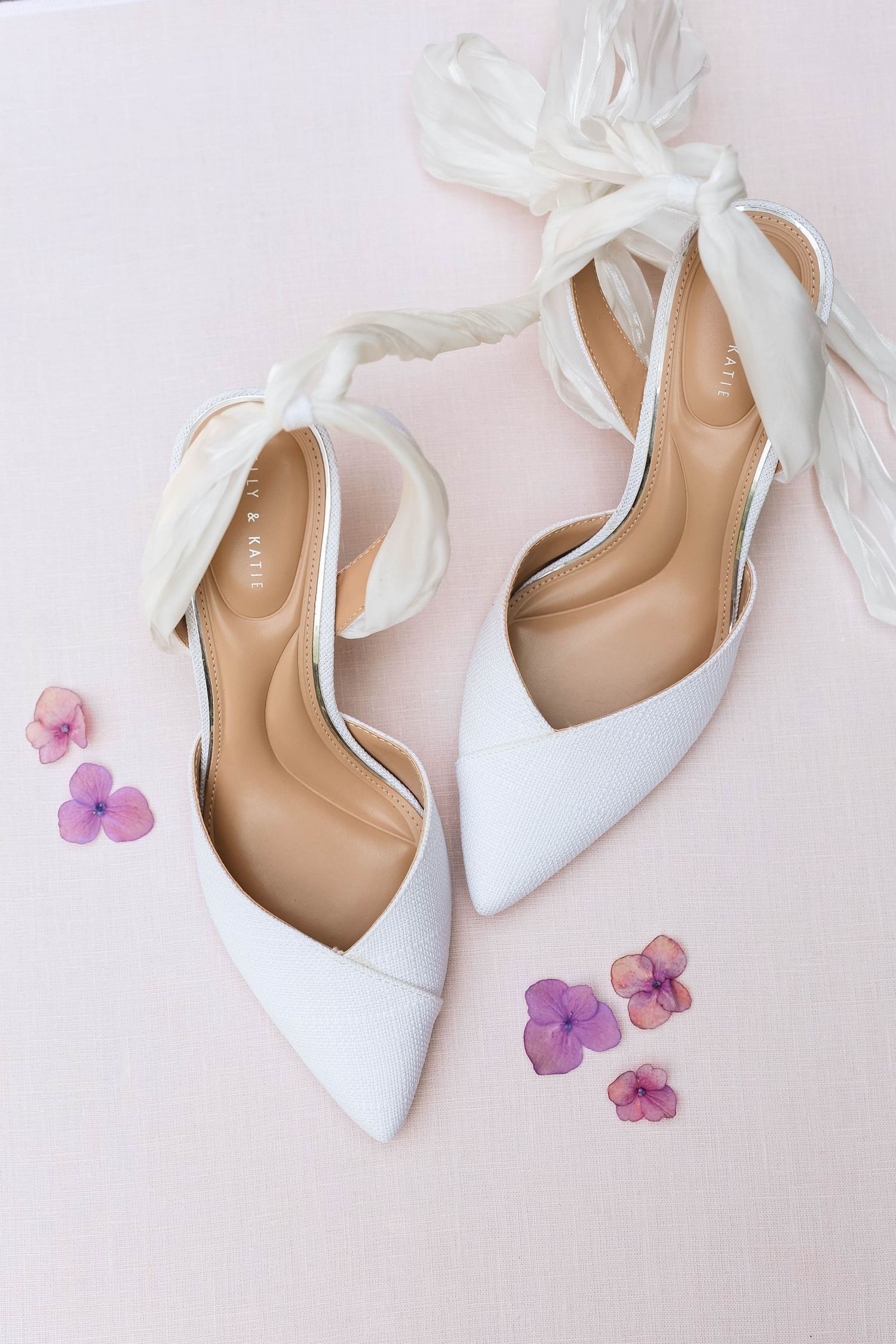 White pointed-toe heels with silk ribbon ankle ties displayed on pink surface with purple hydrangea petals.