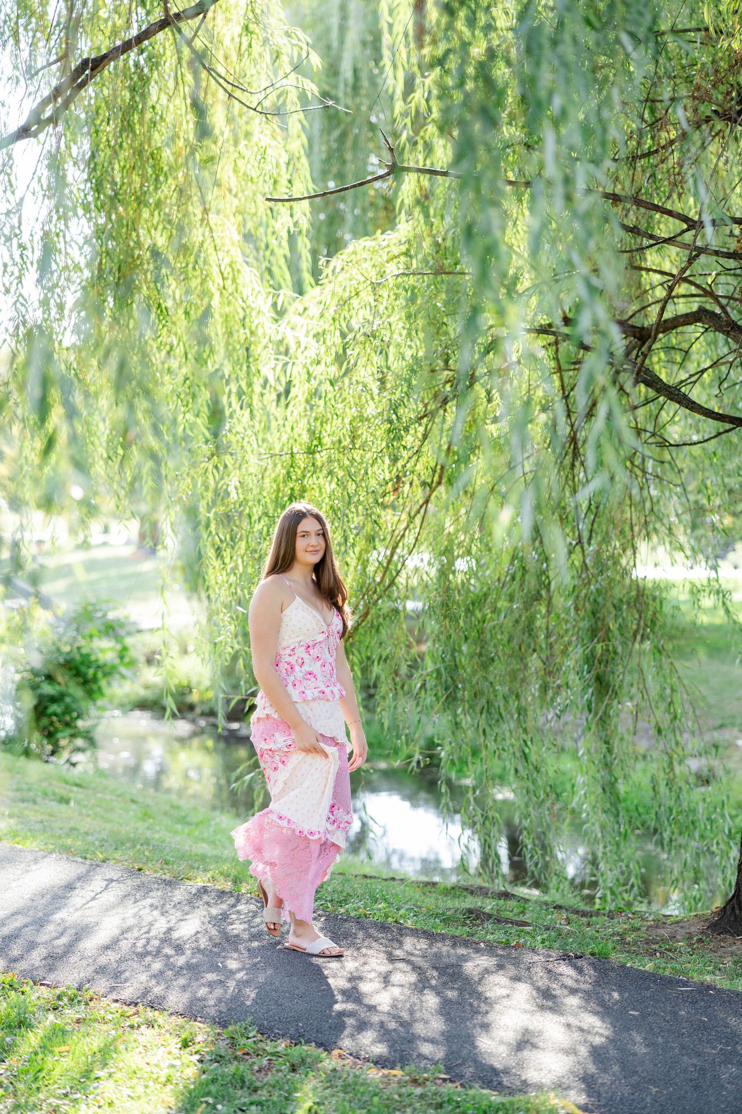 Woman in pink floral dress standing under weeping willow tree by a pond on a sunny day.