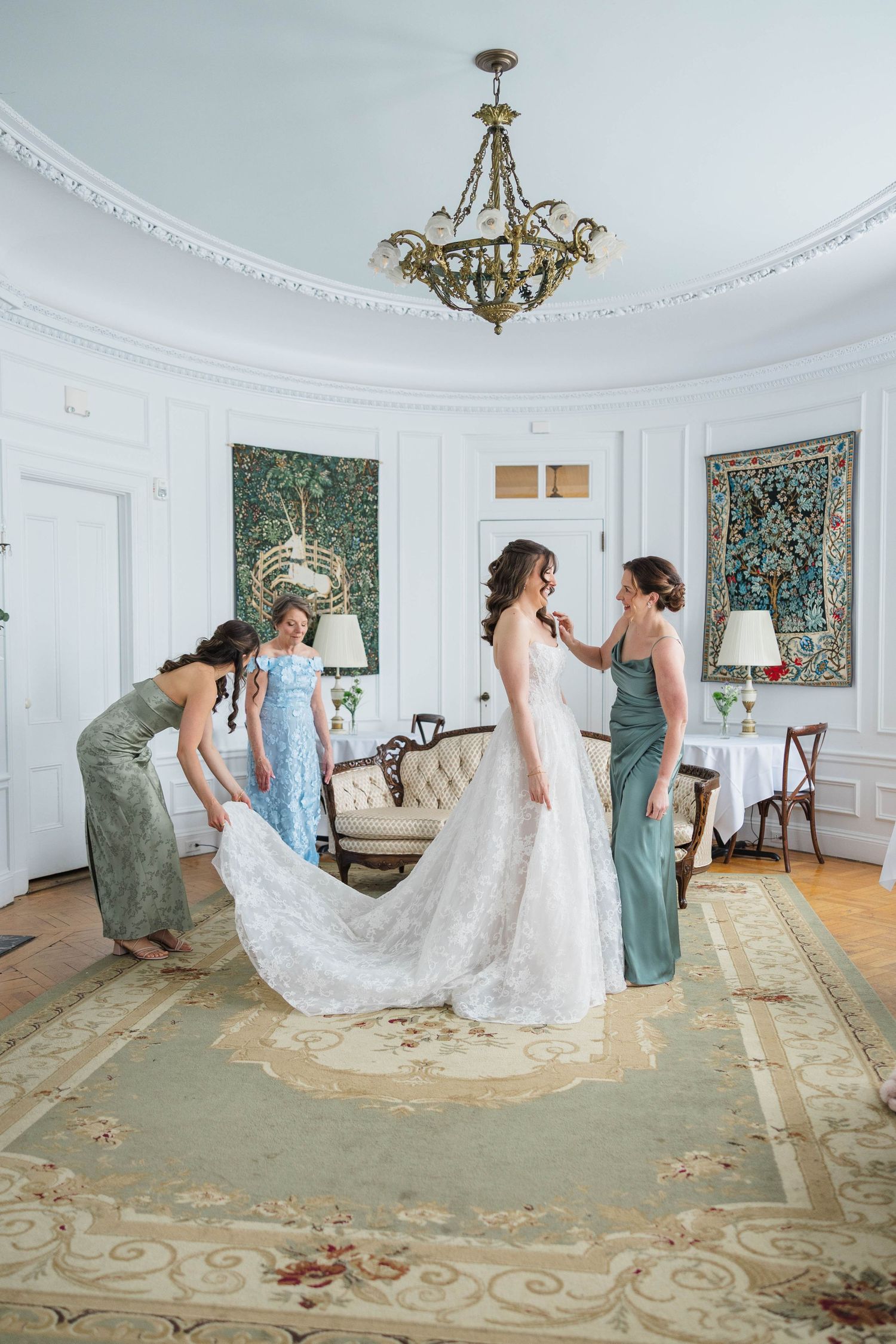 Bride in white lace gown assisted by bridesmaids in teal dresses in an elegant room with tapestries and chandelier.