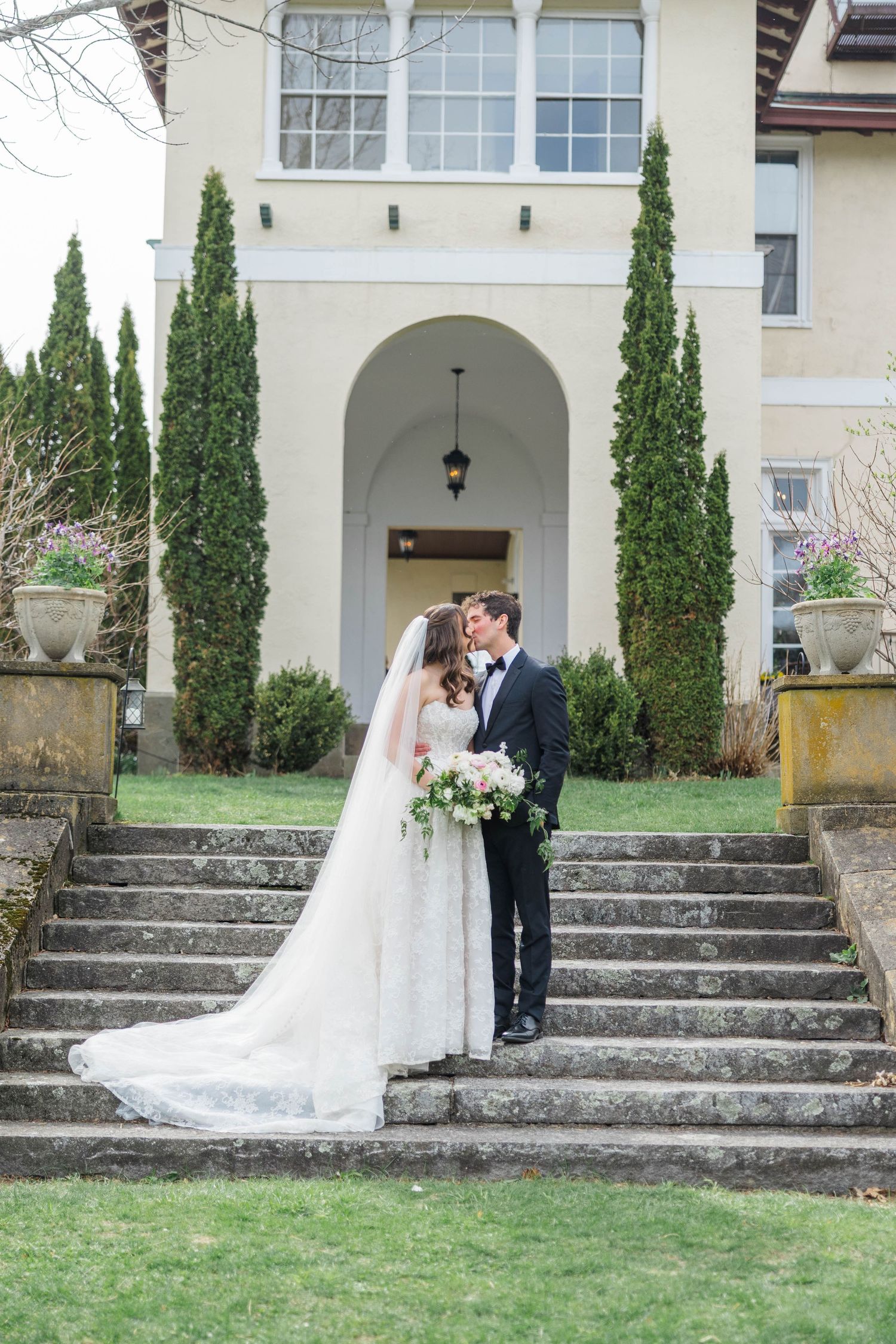Bride and groom share a kiss on stone steps before an elegant villa with tall cypress trees and lush greenery.