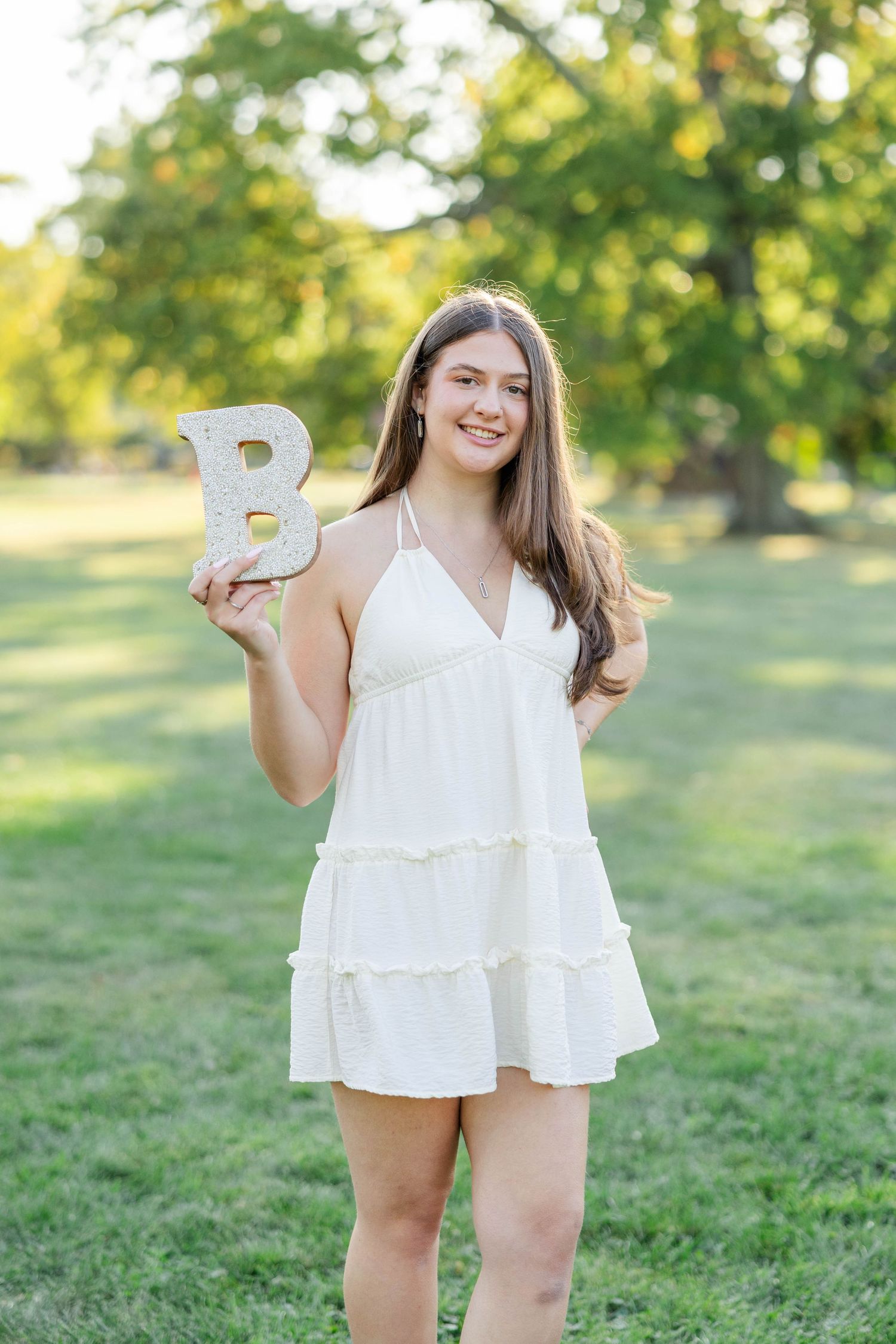 Someone in a white tiered summer dress holds up a decorative letter B while standing in a sunny outdoor setting.