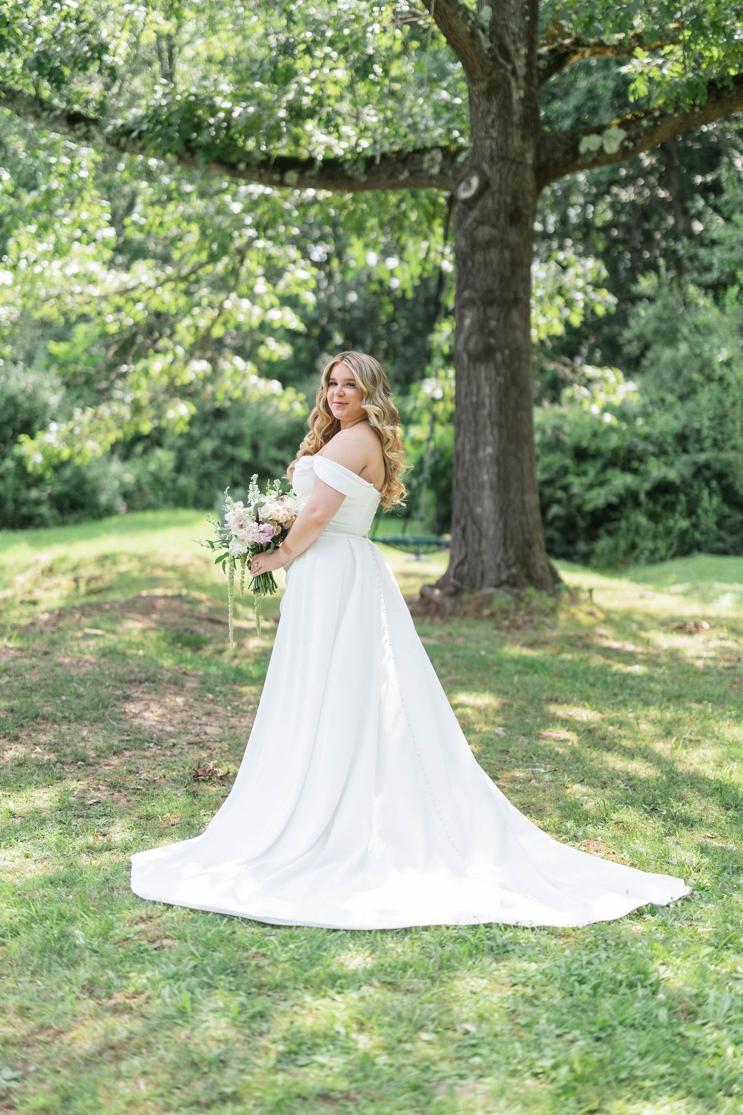 Bride in off-shoulder white gown with train holds bouquet outdoors under large tree on lush green lawn.