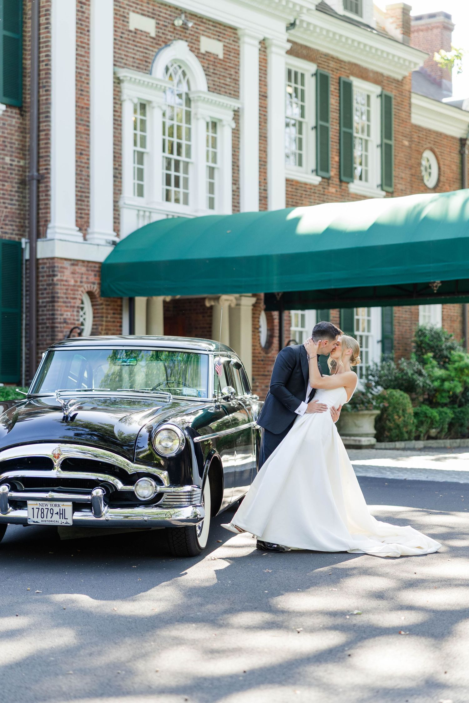 Classic black vintage car parked in front of elegant brick mansion with white columns and green awnings.