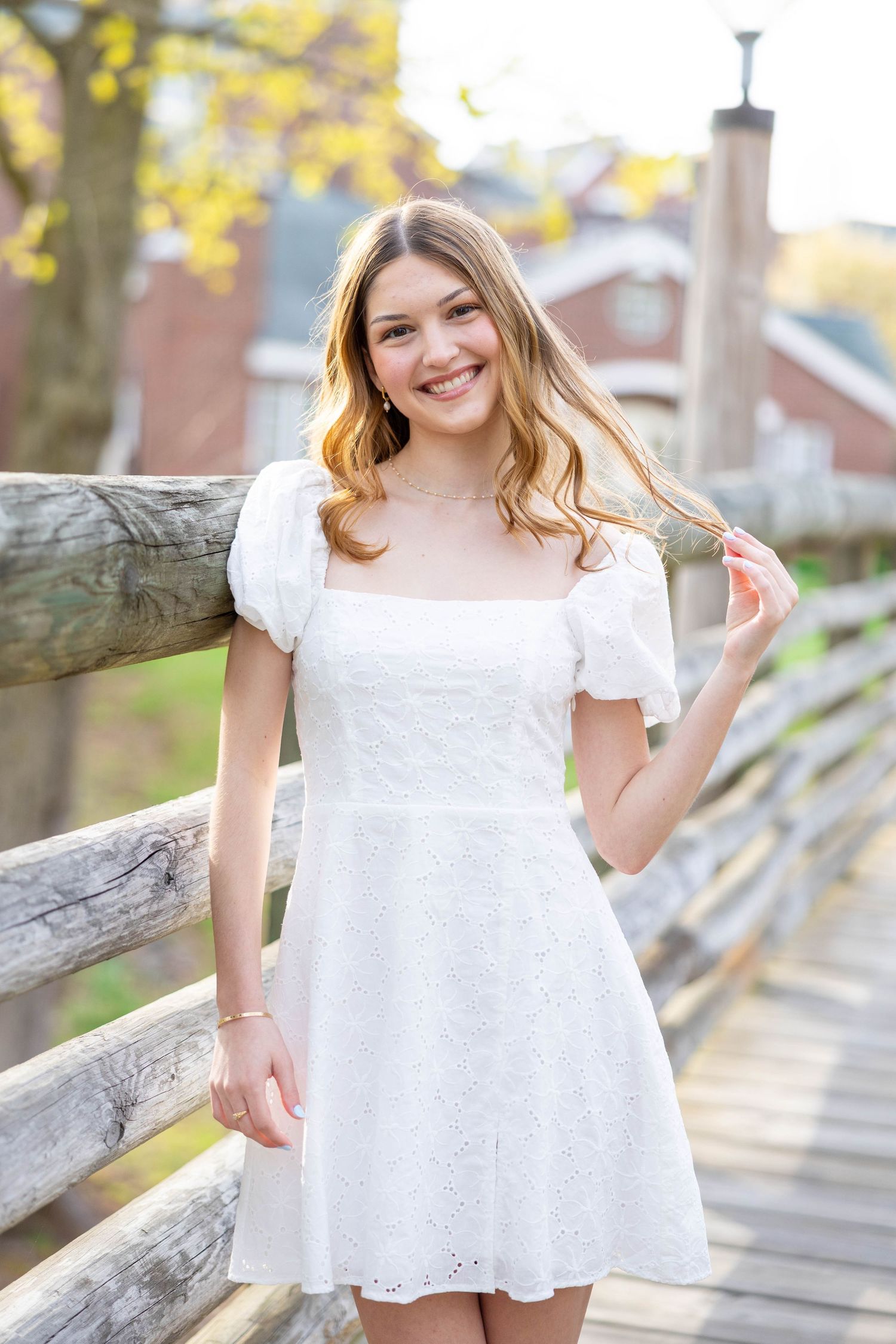 A person in a white eyelet lace dress poses outdoors against a rustic wooden fence in warm sunlight.