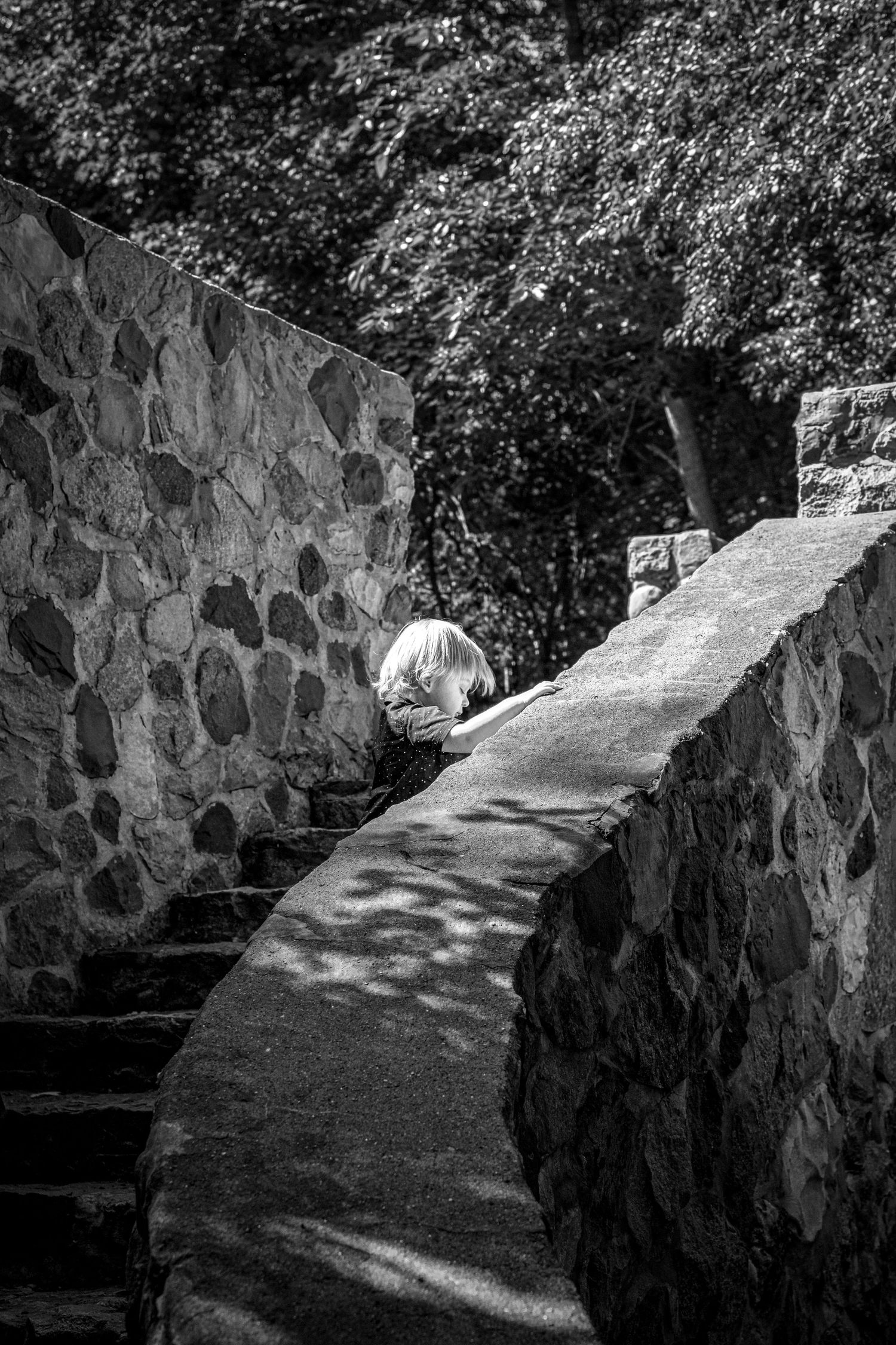 A boy walks up a curved stone wall with steps that create shadows on a pathway surrounded by lush greenery in black and white.