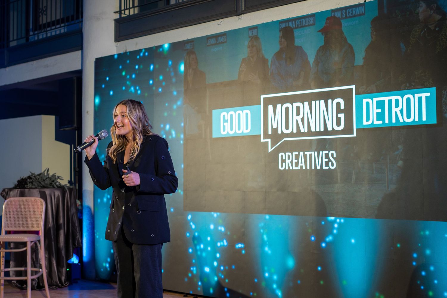 A speaker presents on stage at Good Morning Detroit Creatives event with blue lighting and signage in the background.