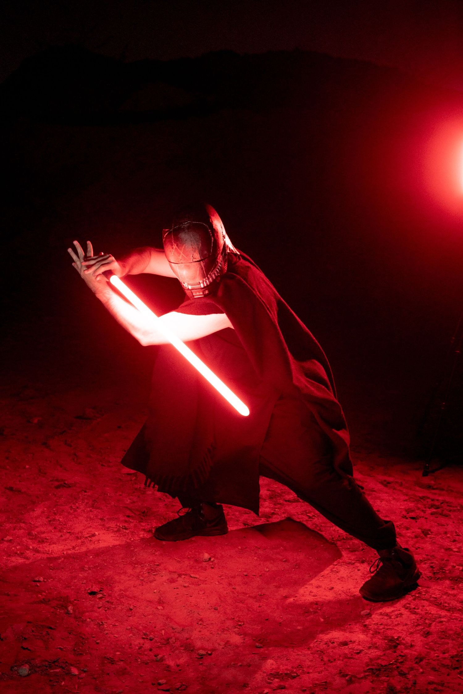 A person in dark clothing performs dramatic pose with glowing red lightsaber against dark background with red lighting effects.