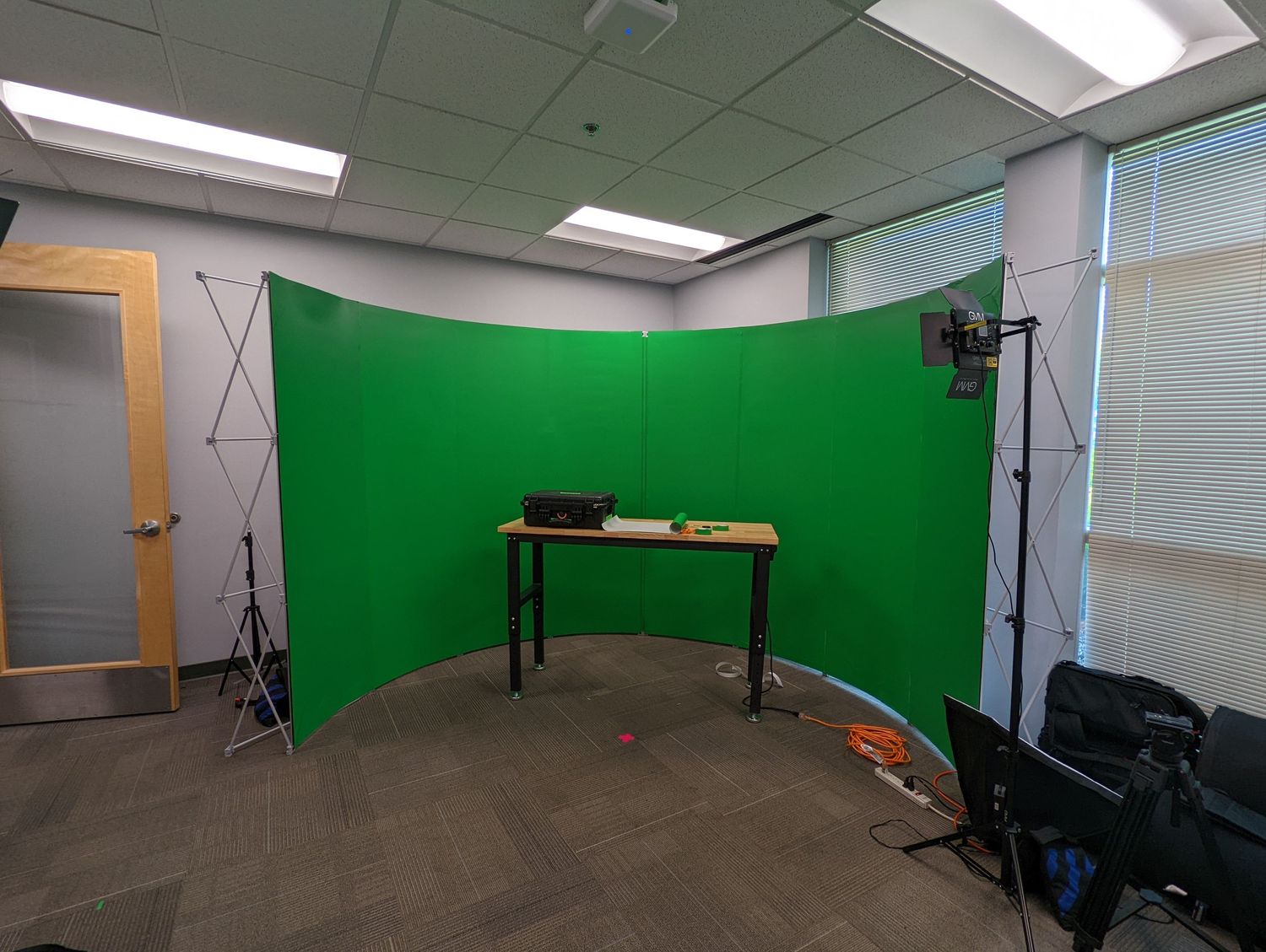 Professional green screen studio setup in corner of office with lighting equipment and desk.