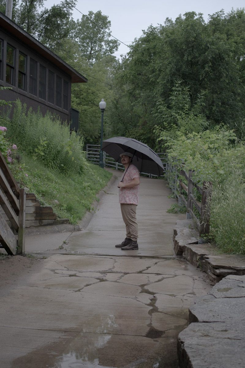 Person with umbrella stands on wet stone path surrounded by lush greenery and a rustic building.