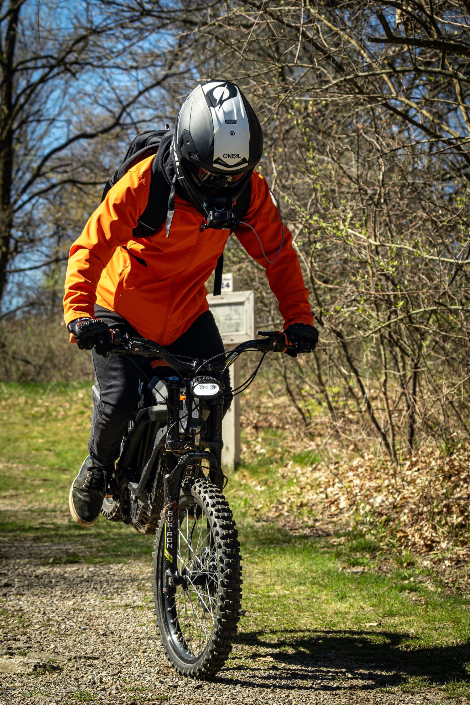 Rider in orange jacket navigating trail on mountain bike through wooded area.