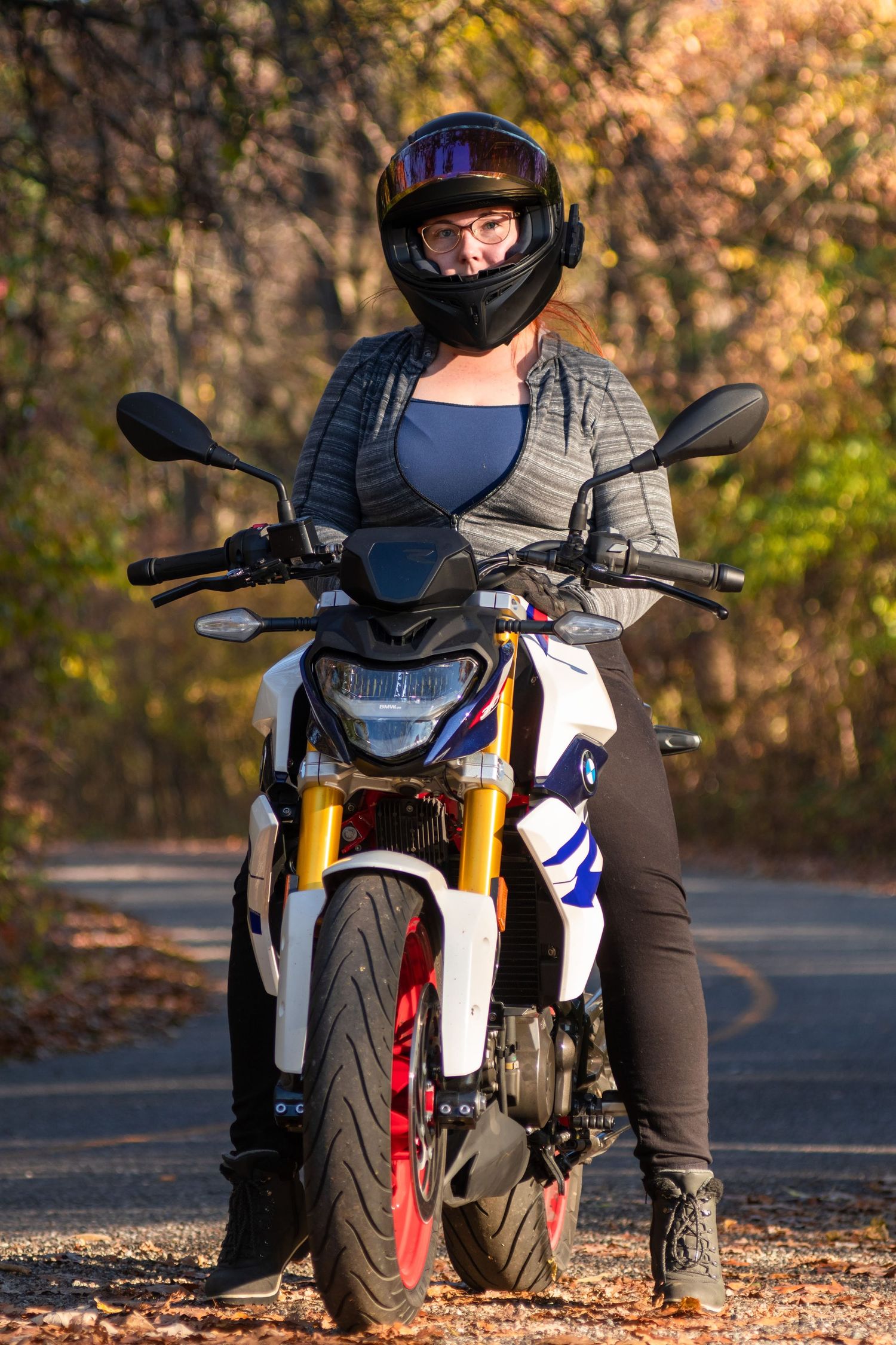 A motorcycle rider in protective gear stands with a BMW G310R bike on an autumn forest road.